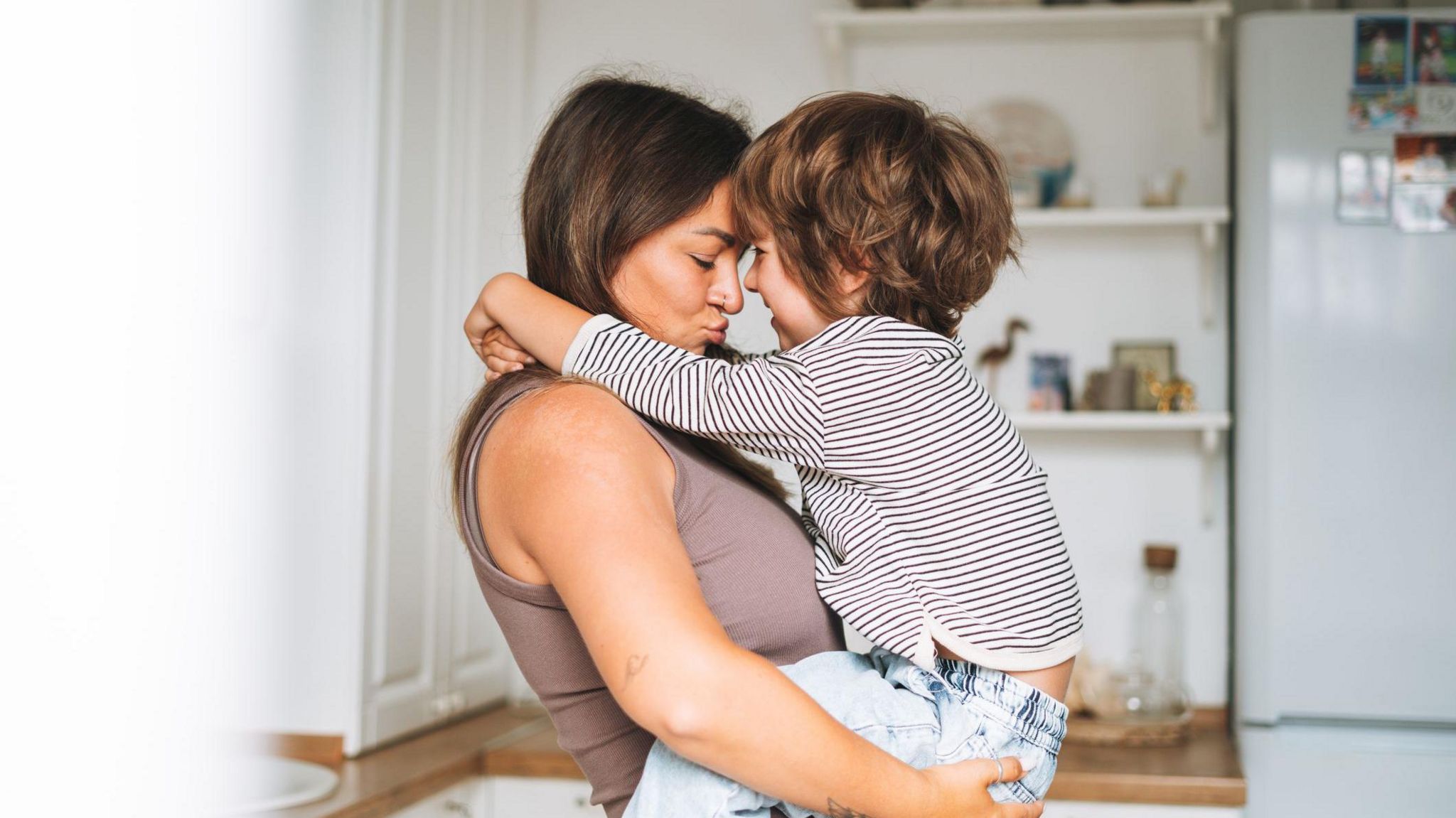 Woman carrying young boy affectionately with a fridge and kitchen shelves seen behind