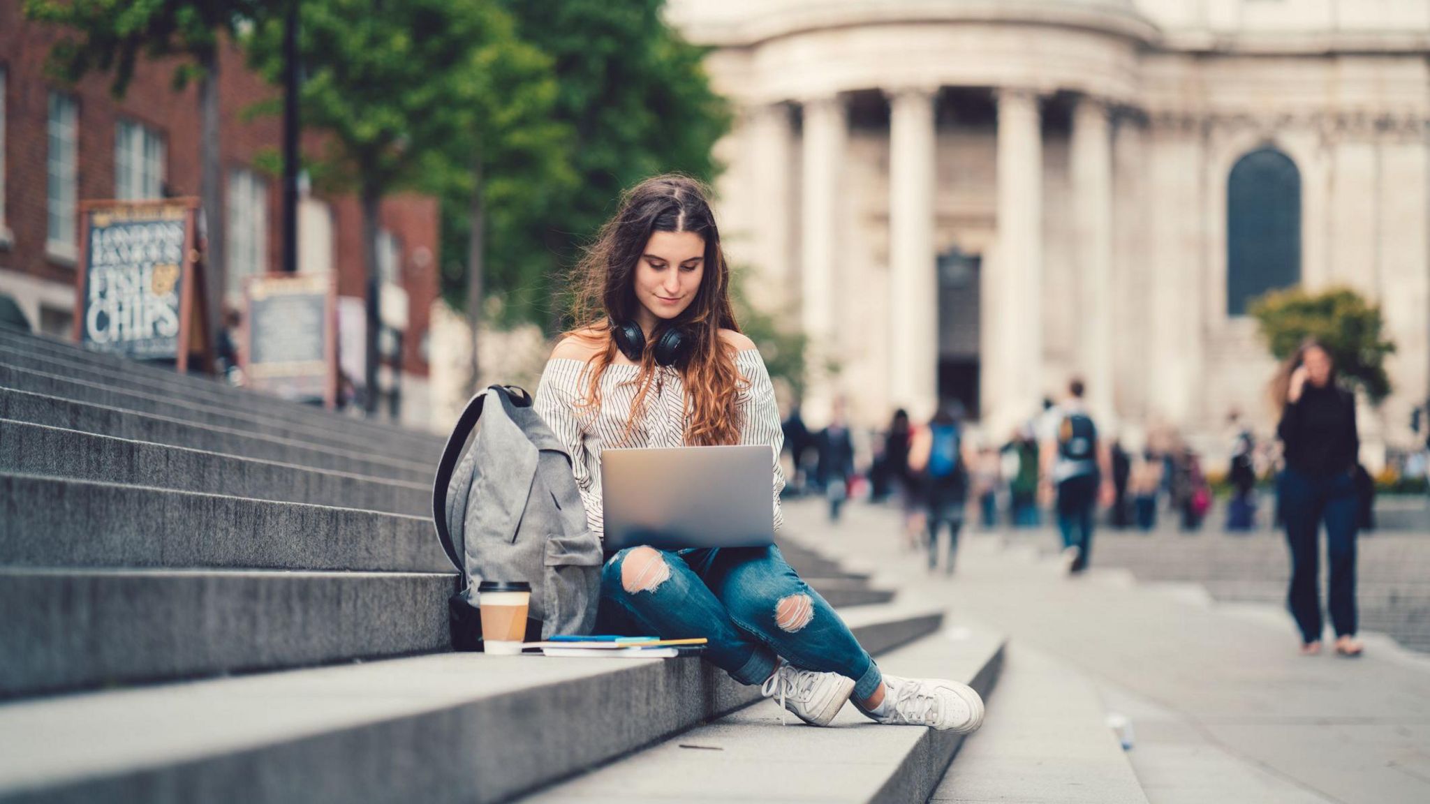 A girl wearing blue jeans and a stripy black and white shirt is sitting on concrete steps on her laptop. She has a bag and a coffee beside her. She is outside an old grey building with people walking past. 