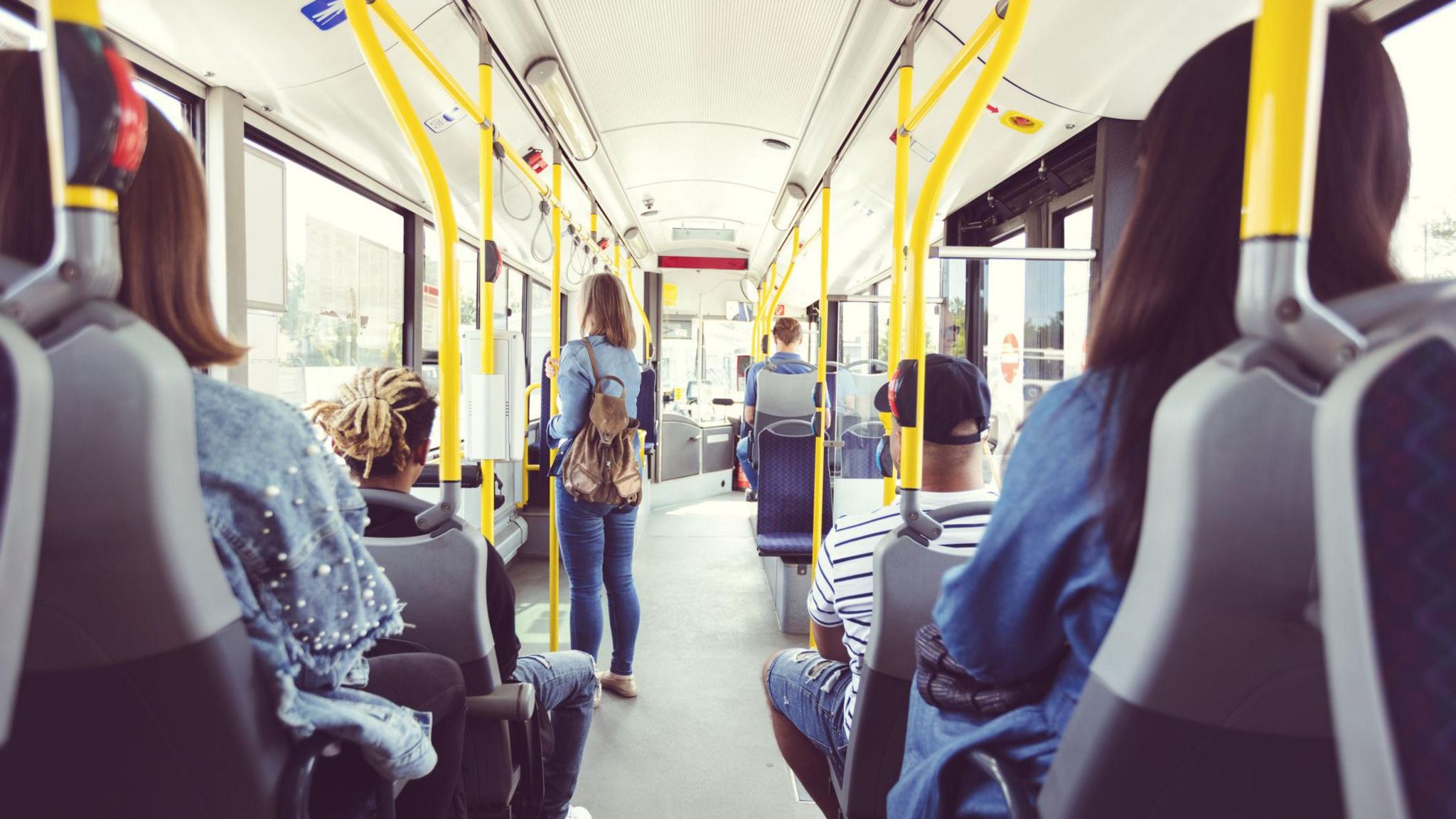 Passengers sitting on a bus seen from behind, with one woman standing