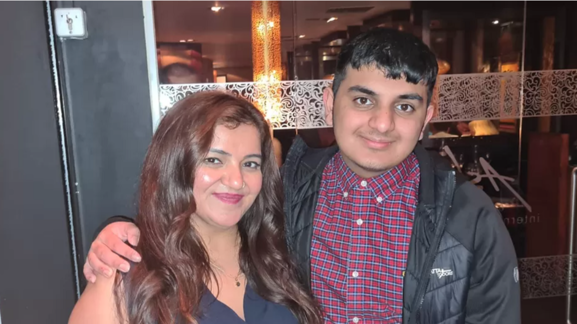 Mum and son smiling and holding each other while looking towards the camera. Mum has long dark reddish hair and son is wearing a blue and red checked shirt with a black jacket on top