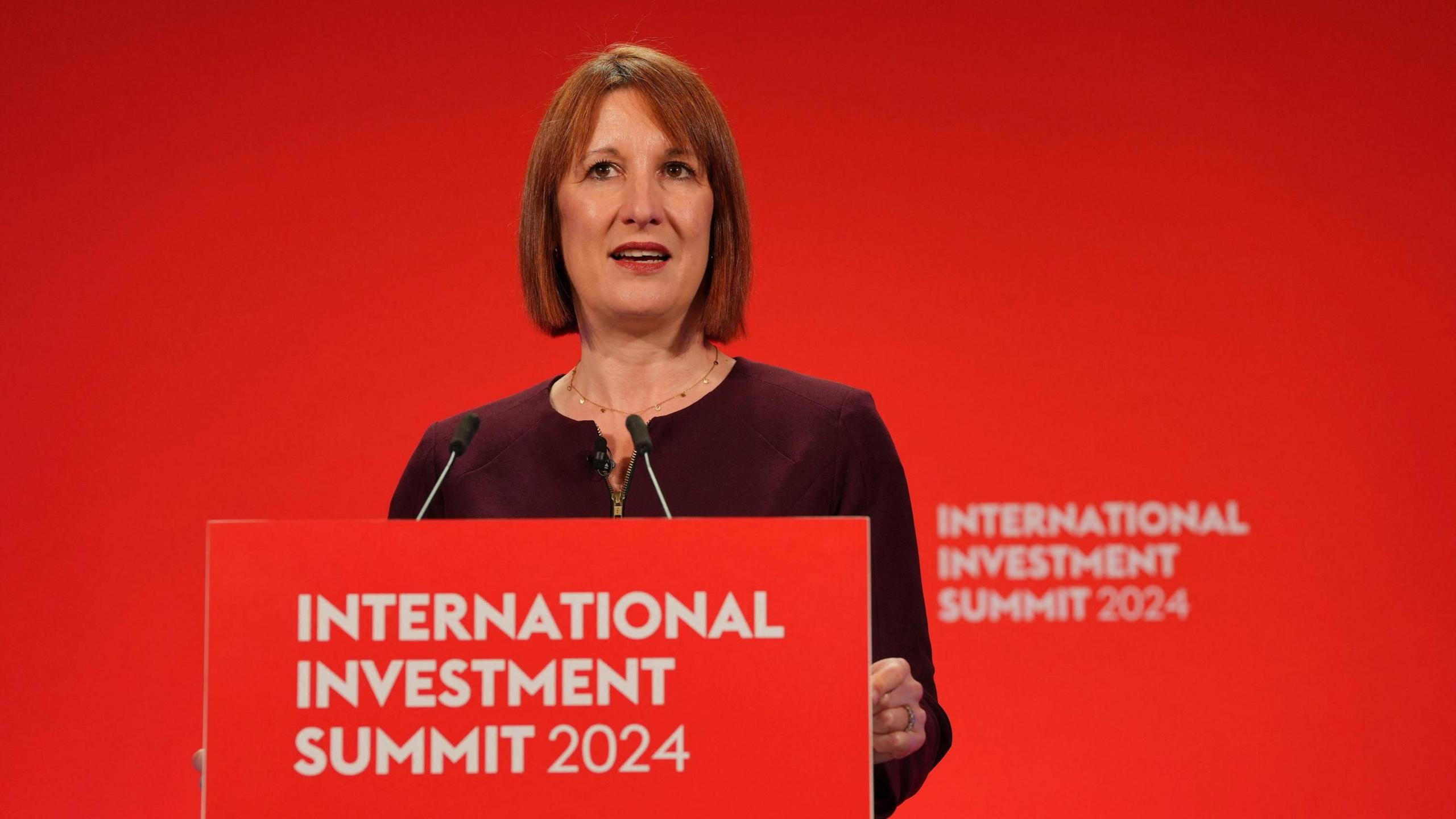 Rachel Reeves wears a maroon dress as she stands at a red podium which reads: International Investment Summit 