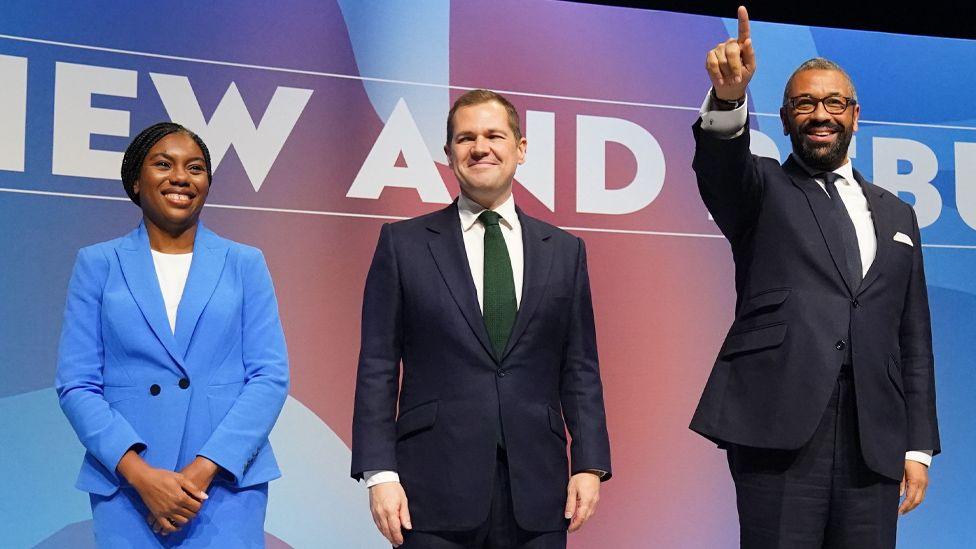 The remaining candidates, Kemi Badenoch in a bright blue suit, Robert Jenrick in a navy blue suit and Cleverly in a navy blue suit, stood next to each other on stage at the Conservative Party conference - all smiling at the crowd.