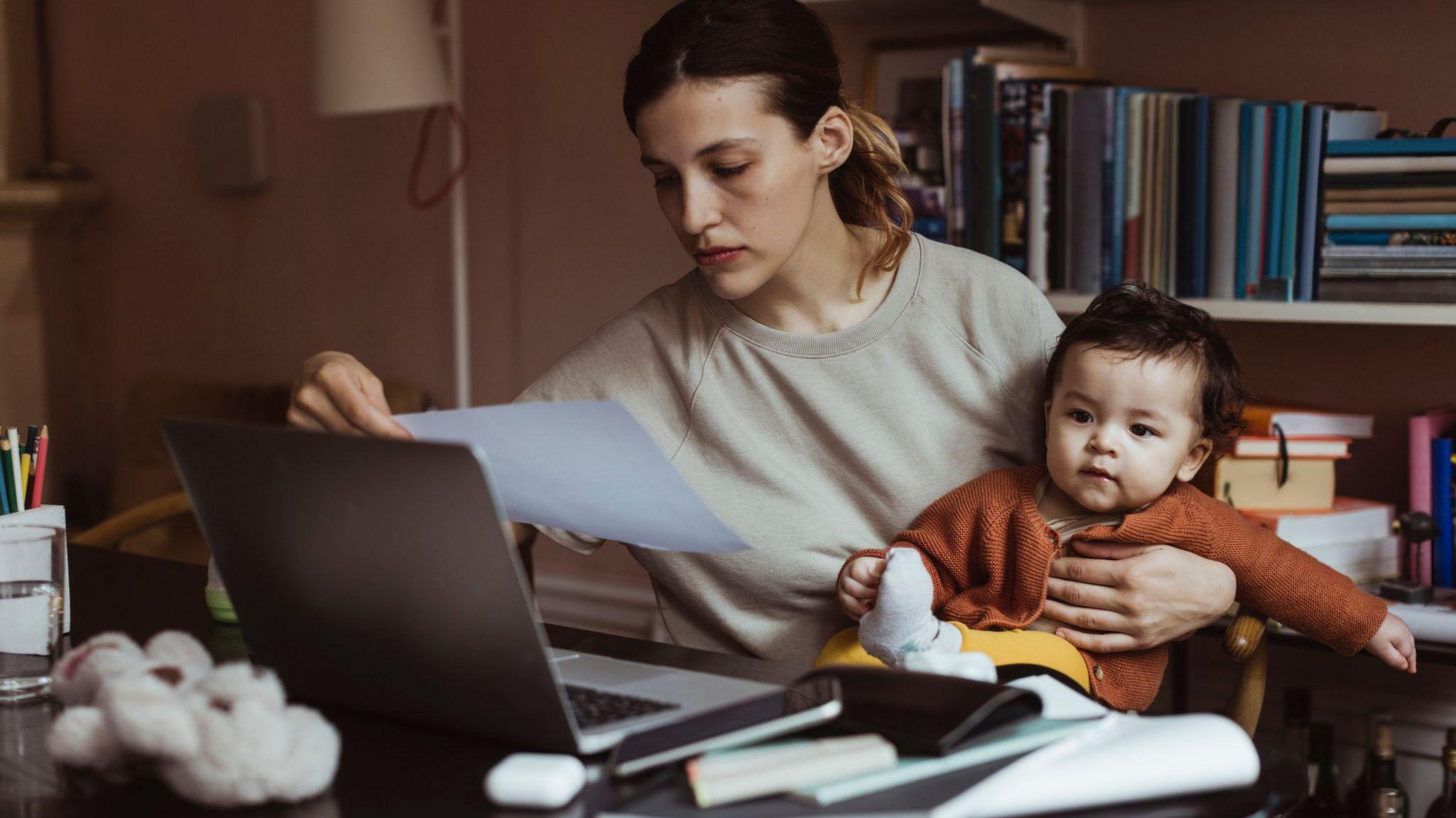 Female professional wearing a grey jumper balances her baby while sitting down at a desk at home where she is reading a document