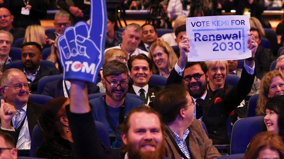 A bearded man in the audience at the Tory conference smiles as he holds up a foam finger