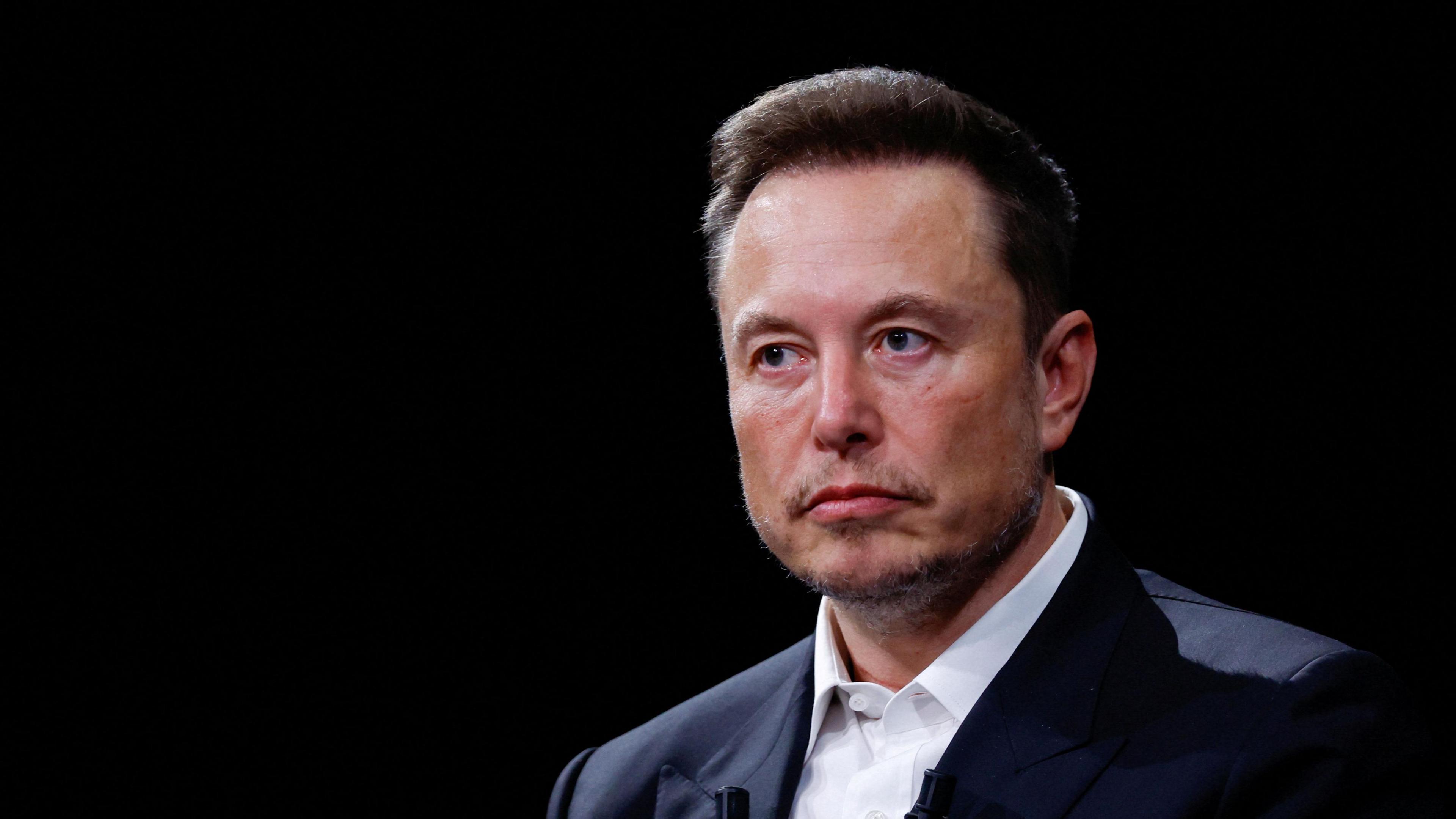 Elon Musk, wearing a black suit jacket and white shirt, sits against a black backdrop on stage at an event.