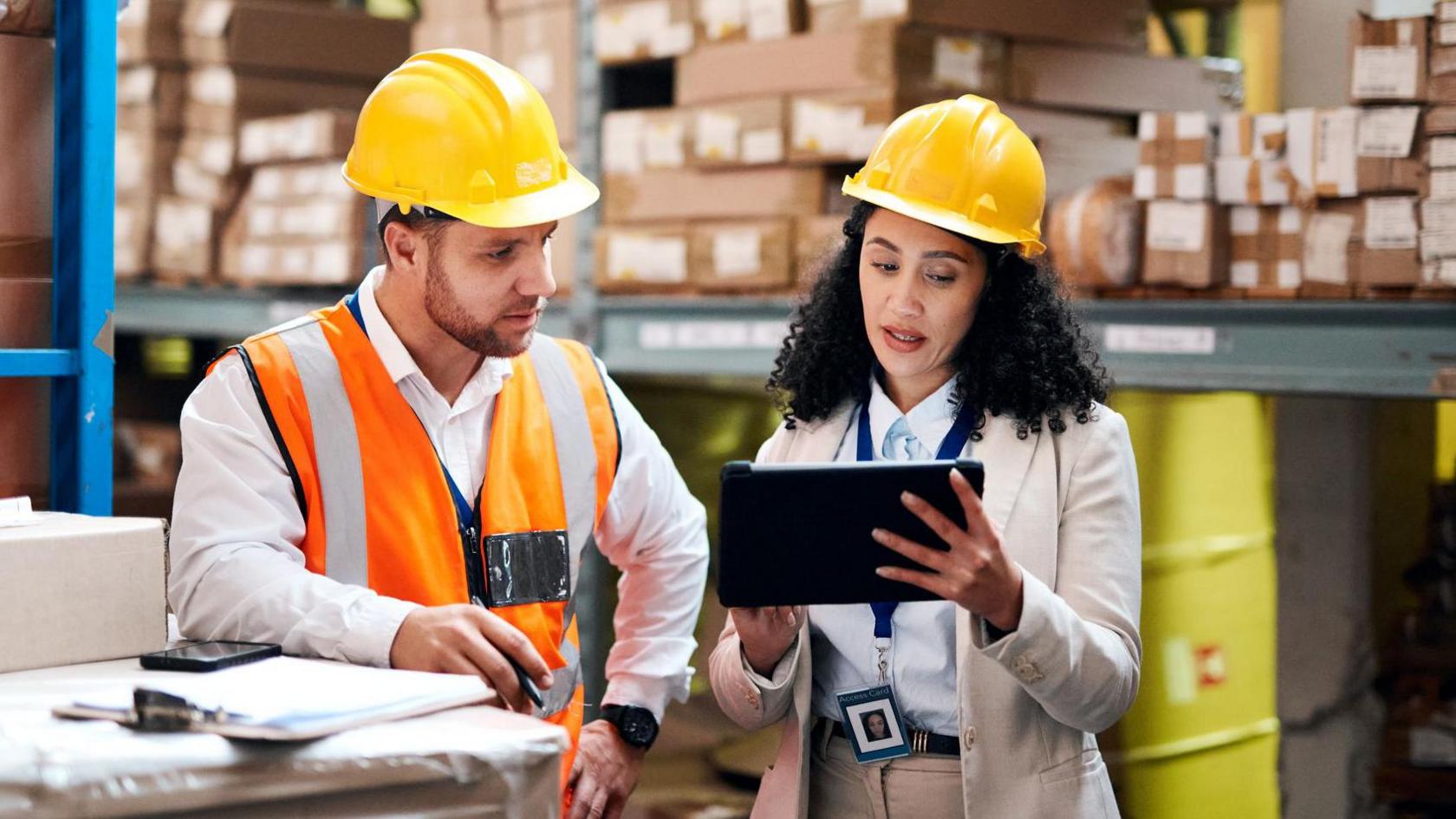 Employees wearing hard hats in a warehouse discussing business while looking at an computer tablet