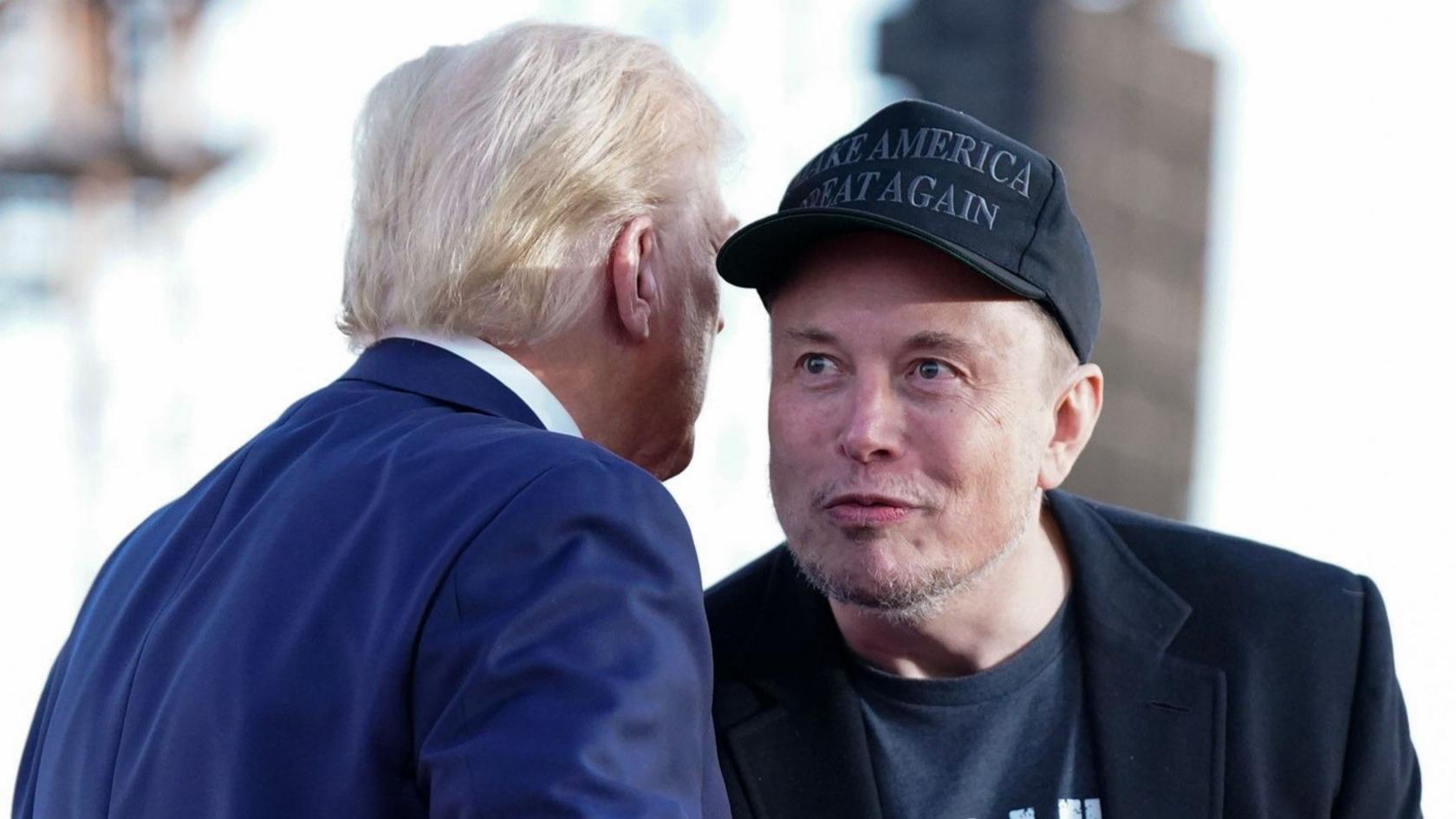 Tesla and SpaceX CEO Elon Musk speaks with former president Donald Trump during a campaign event in Pennsylvania. Musk wears a black 'Make America Great Again' hat and black blazer while Trump wears a blue suit.