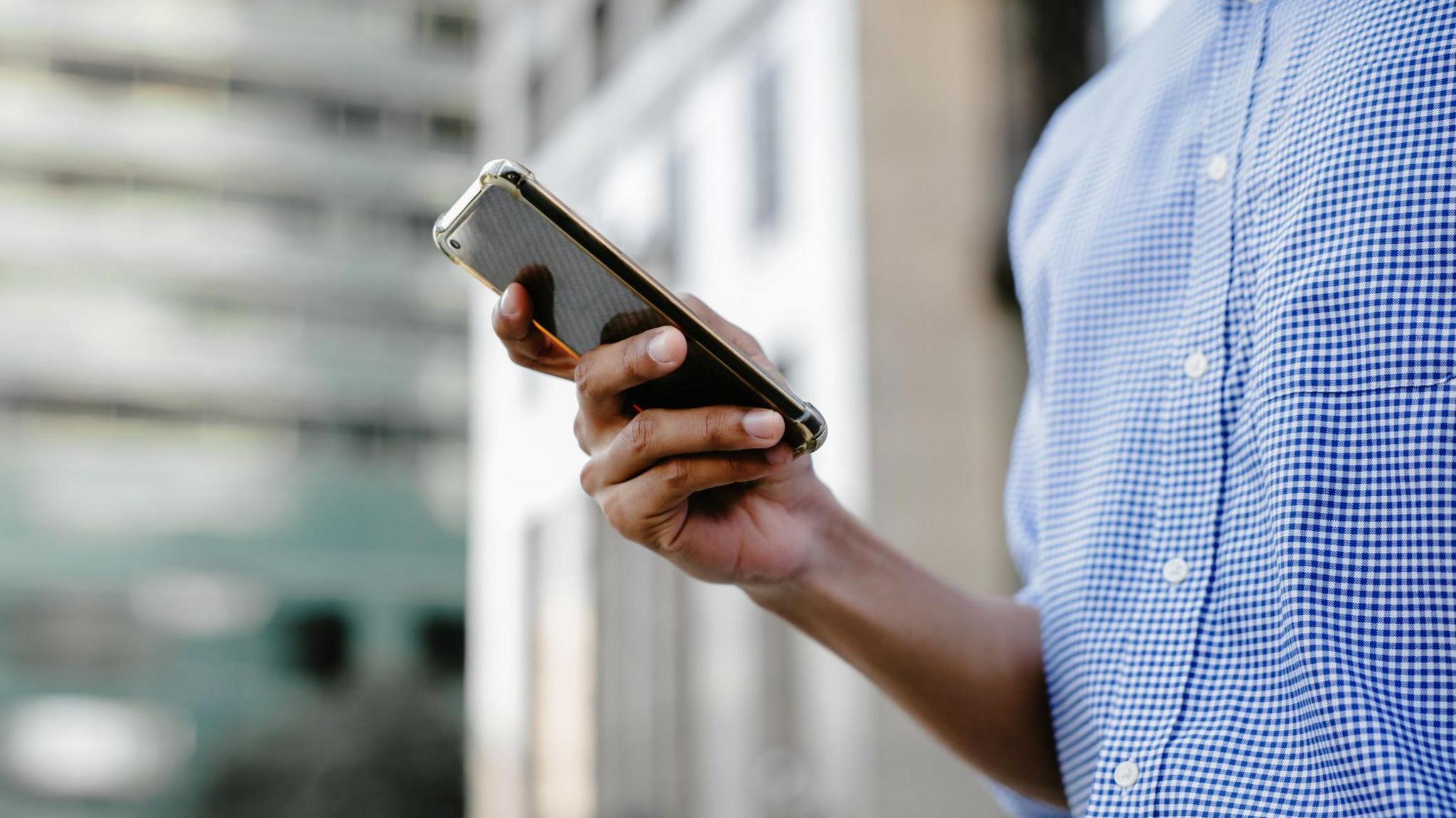 A man in a blue and white checked shirt scrolls on his smartphone, with blurred buildings displayed in the background.