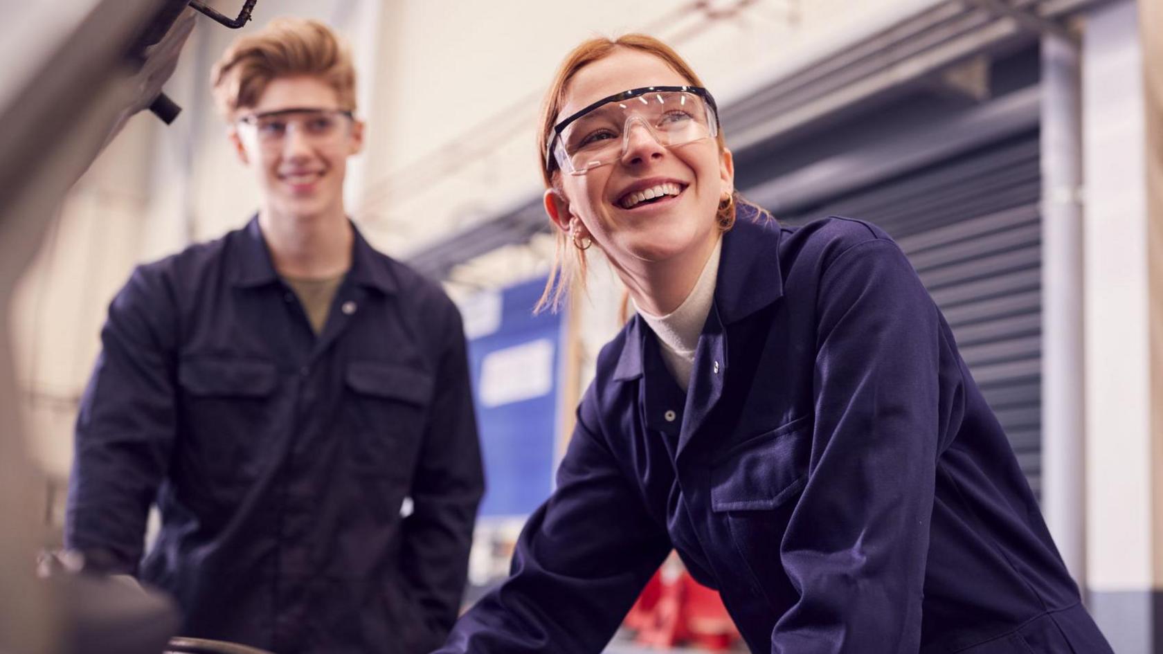 Two young apprentices, one male and one female, smile with looking at a car engine while wearing blue jumpsuits and glasses