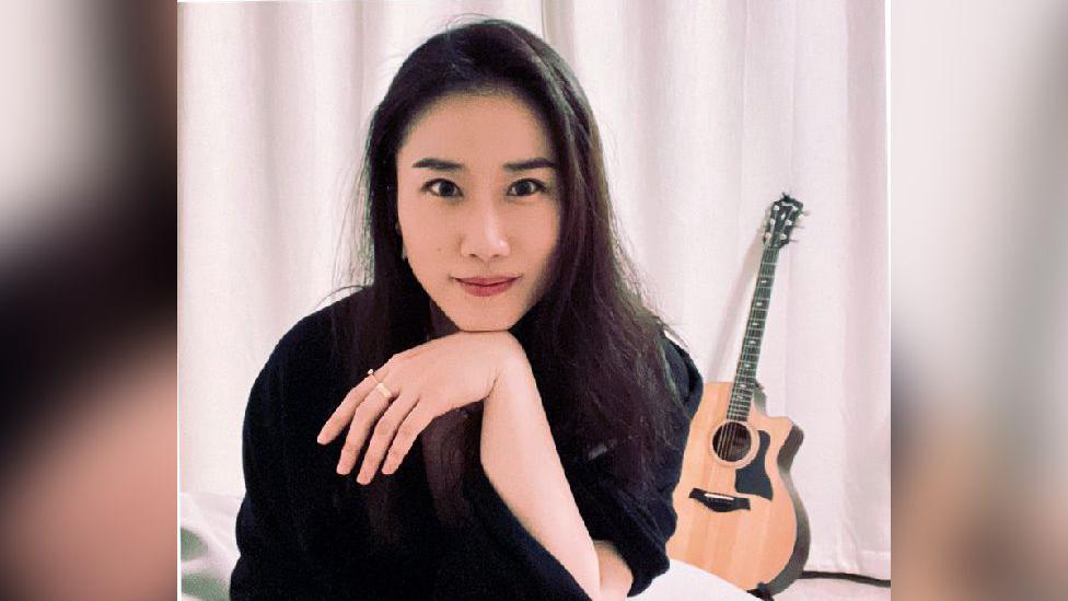 A woman dressed in black sits in front of a white curtain, her chin resting on the wrist of her hand. An acoustic guitar sits on a stand in the background.