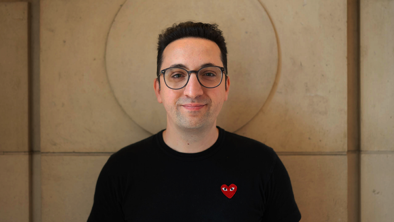 A man with short black hair and glasses and a black t-shirt stands against a sand-coloured wall. His head is perfectly framed by a circular indentation carved into the large stone block.