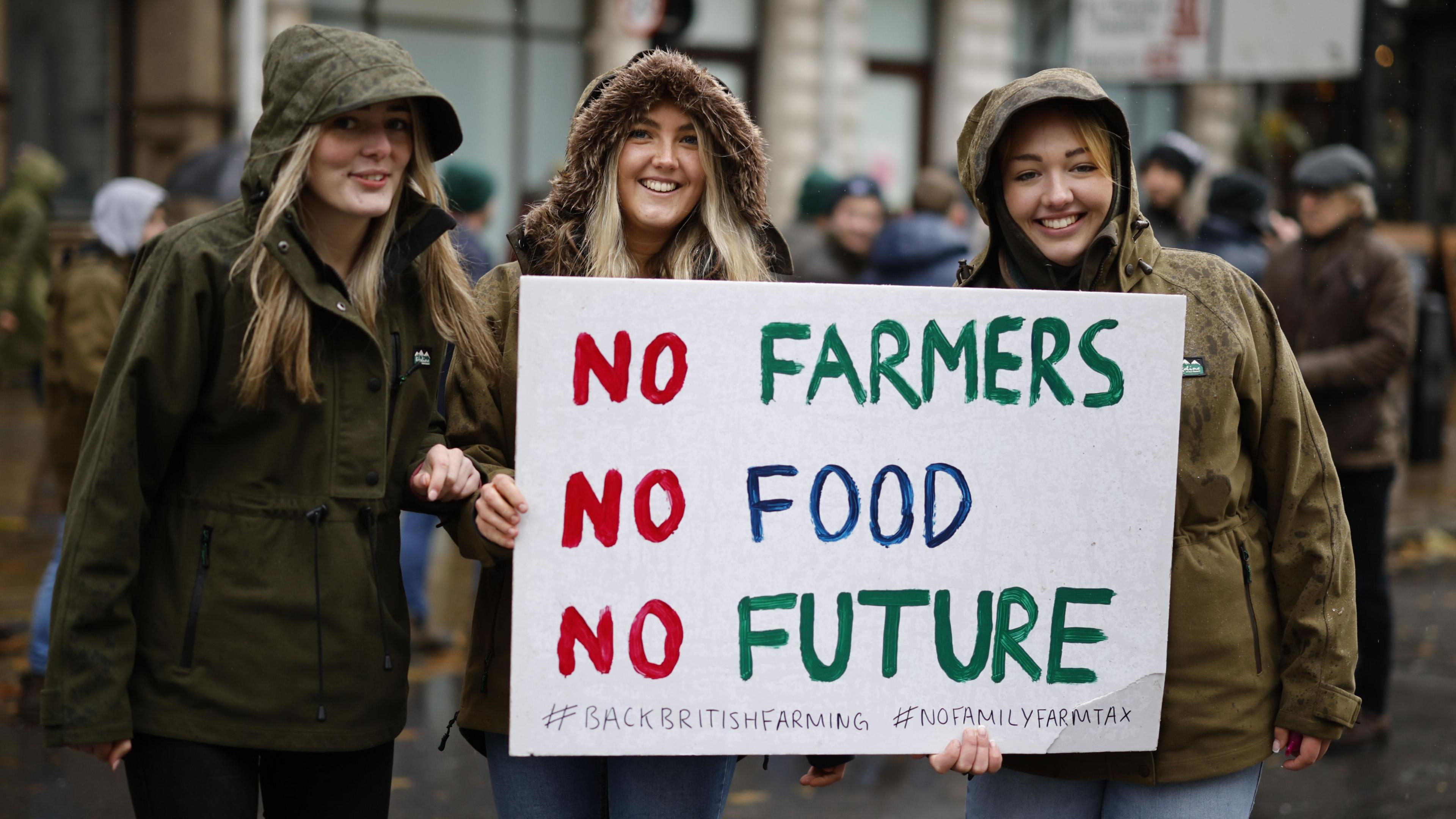 Three women wearing coats with their hoods up in the rain hold a sign at the protest which reads 'no farmers, no food, no future'