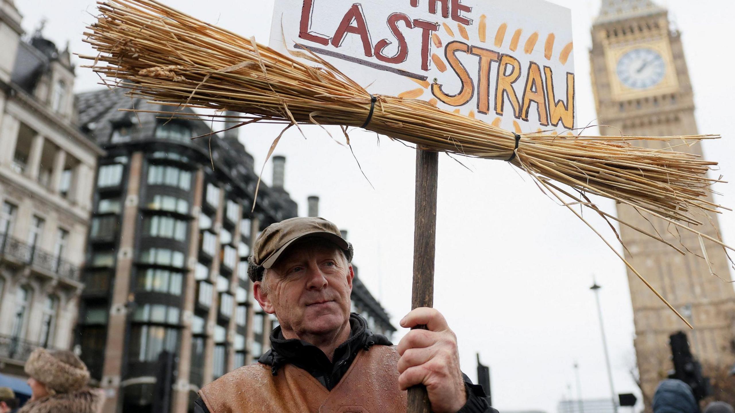 A man taking part in the protest wears a leather waistcoat and flat cap. He holds a sign which reads 'the last straw' and which has straw attached to it. In the background is Big Ben