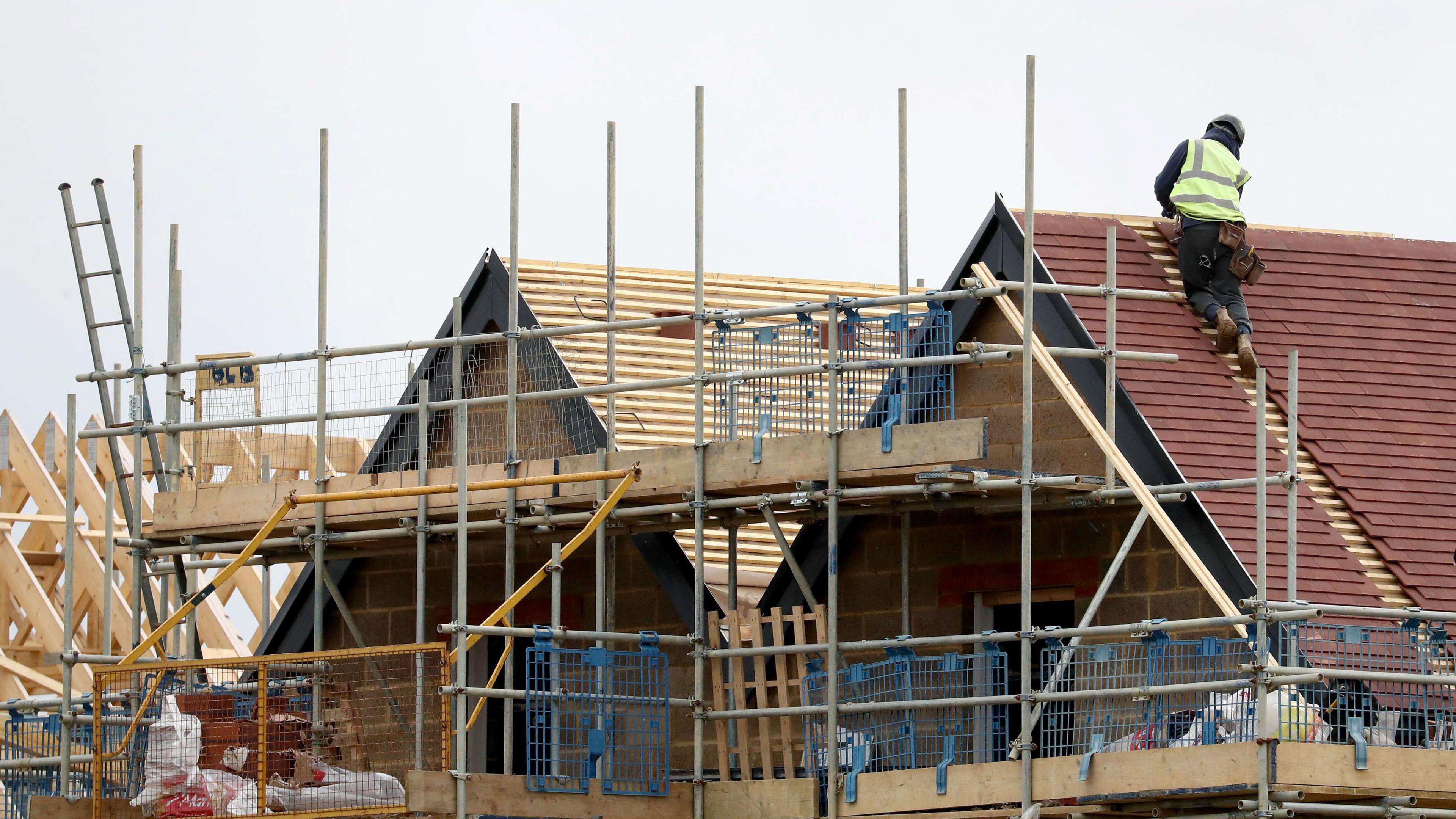 A construction site showing a house in progress, with scaffolding surrounding the structure and a worker in a high-visibility vest installing roof tiles on the sloped roof. Wooden beams and other construction materials are visible in the foreground and background