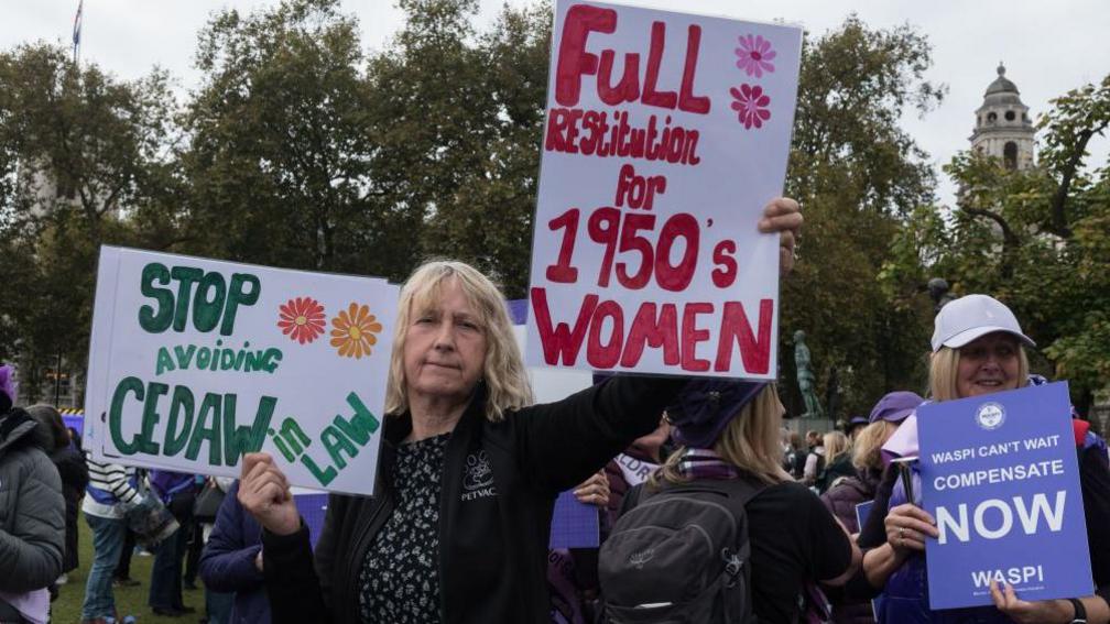 A woman at the protest holds two signs. One reading "stop avoiding CEDAW in law" and another says "full restitution for 1950s women". 