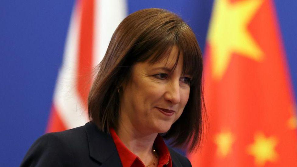 UK Chancellor Rachel Reeves dressed in a black jacket and red shirt standing in front of a backdrop of two flags representing China and the UK