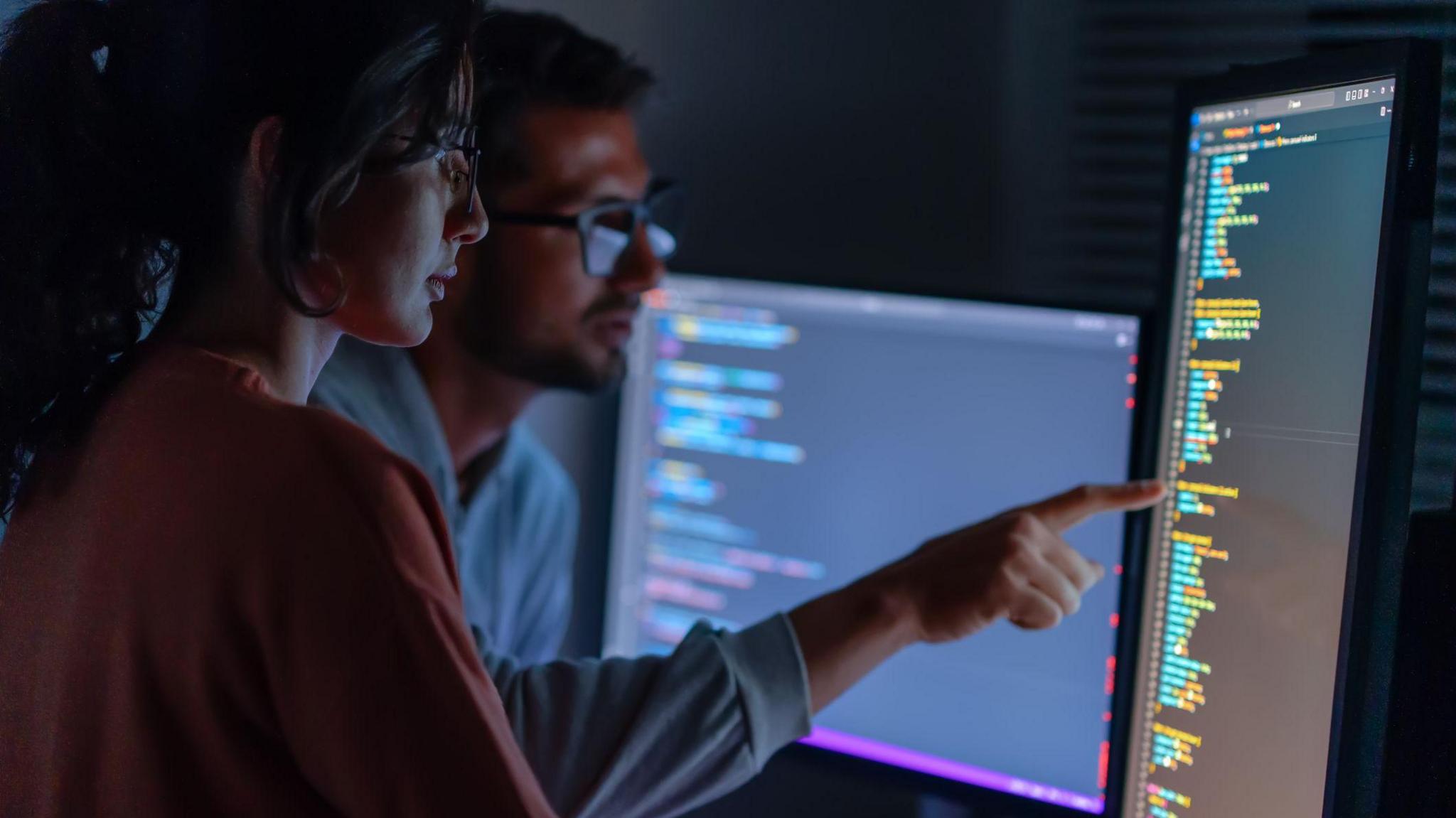 A woman and a man look at a computer screen containing lines of code. The man is pointing at one of the lines on the screen.