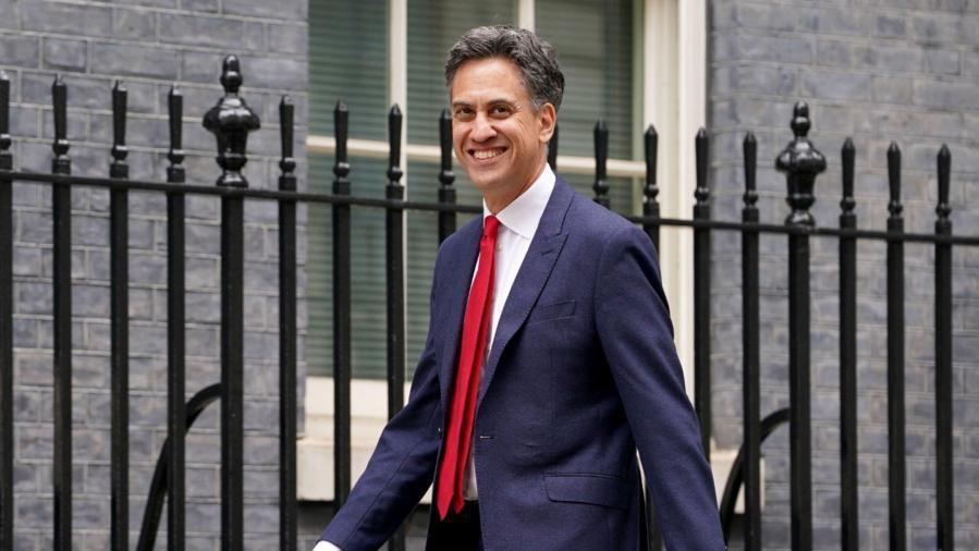 Ed Miliband in a suit and red tie walking beside a fence 