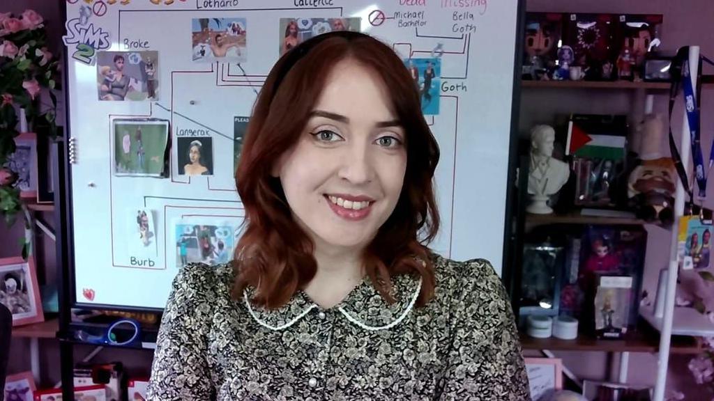 A woman with dark red hair wears a floral print dress as she stands, smiling, in front of a white board and some shelves cluttered with ornaments. The whiteboard has printouts of screenshots from The Sims stuck to it, with lines and notes in black marker linking them up.