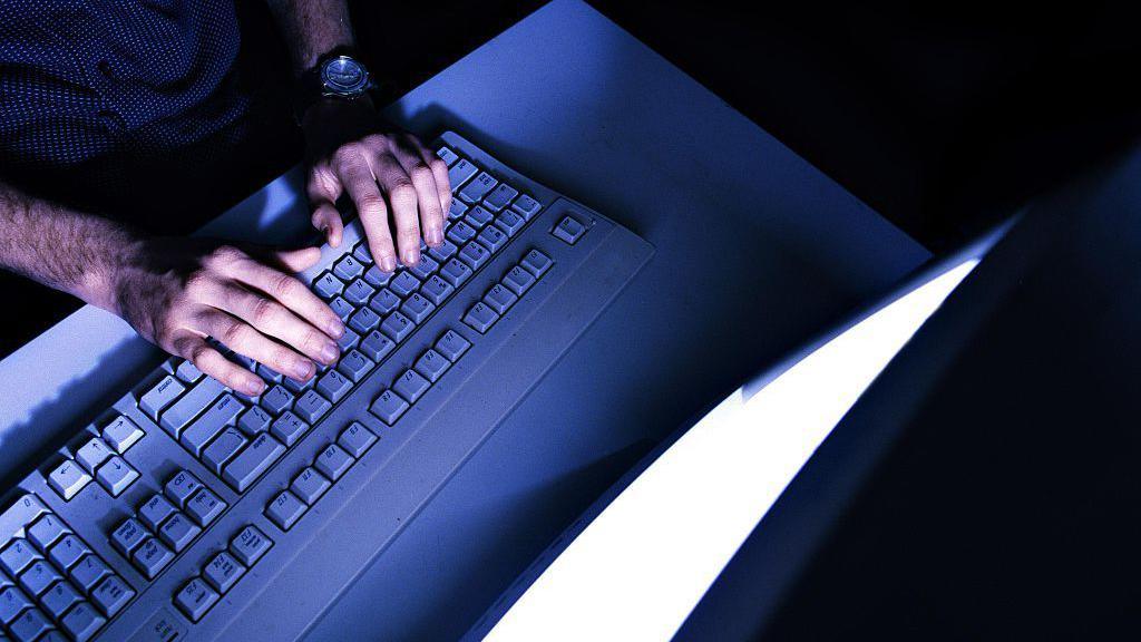 A man places his hand on a computer keyboard while looking at a screen, his face is not in shot