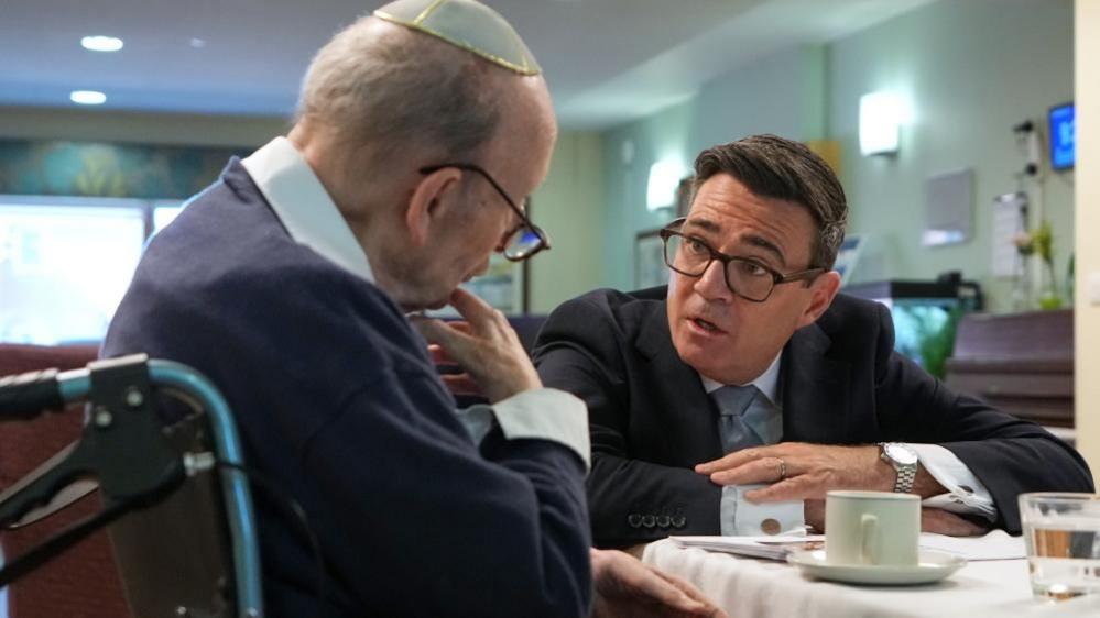 On the left is a man sat in a wheelchair. He is wearing a blue jumper, a white shirt, glasses and a kippah. To his right is Andy Burnham, who is also sat down with his arms placed on a table. Burnham is wearing a dark-coloured suit jacket, white shirt and a tie. The two men are in discussion. This is taking place in a care home.