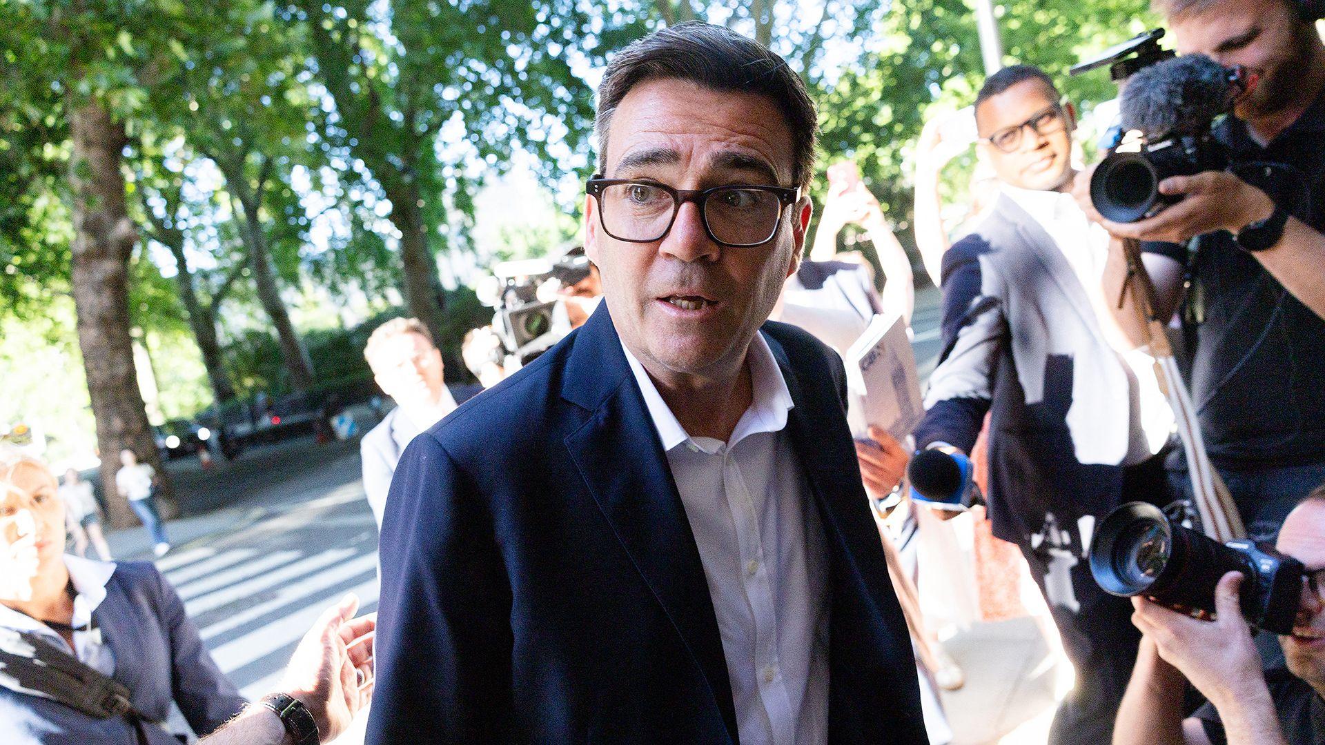 Andy Burnham looks to the left of frame while wearing a suit and shirt and surrounded by cameras and reporters, as he arrives at Millbank on 2 July
