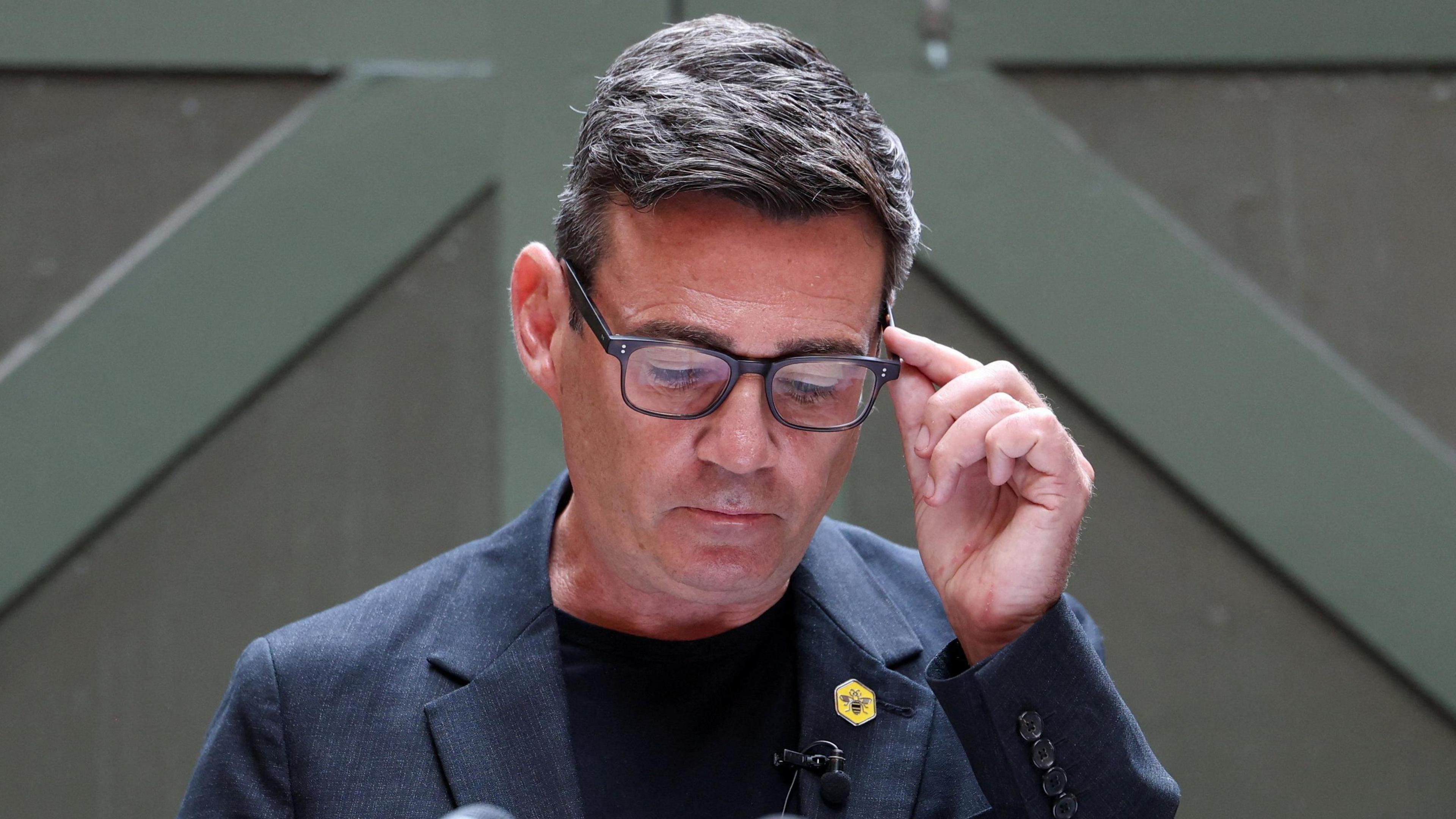 Andy Burnham looks down, holding the side of his glasses. He wears a blue jacket, blue top and a yellow badge with a bee symbol on it.