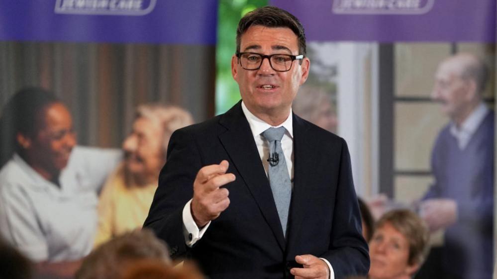 Andy Burnham is wearing a dark-coloured jacket, a white shirt, a tie and glasses. He is speaking to an audience who are not pictured.