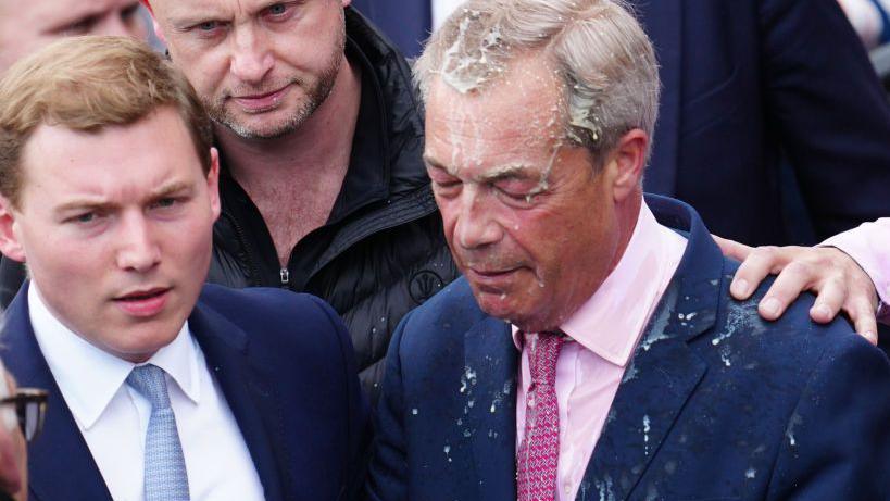 Reform UK party leader Nigel Farage reacts after a woman threw a drink over him. On his left is a second man, dressed in a suit with a white shirt and blue tie.