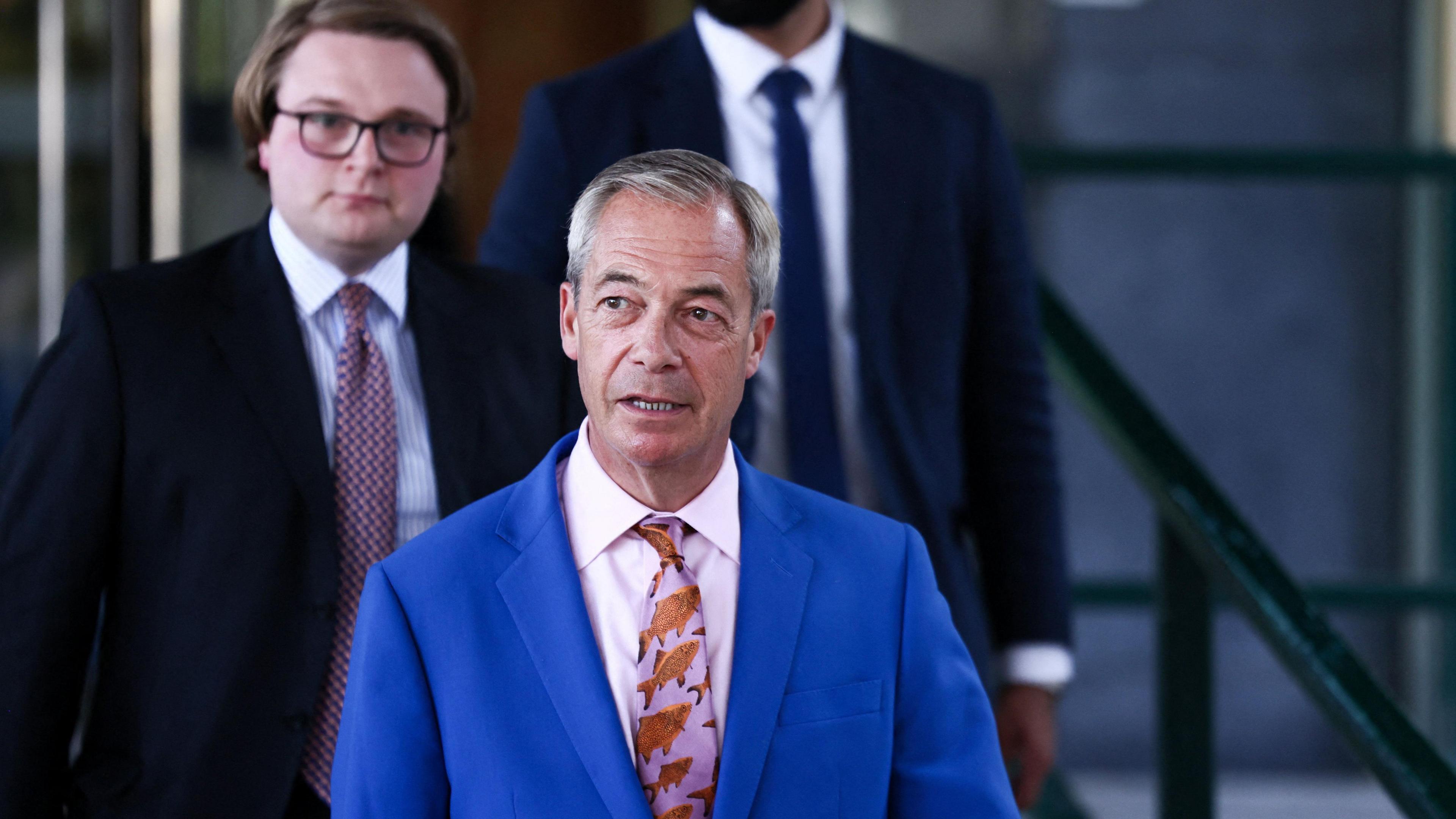Nigel Farage walking outside, wearing a blue suit with a pink tie, decorated with a fish pattern. There are two men behind him, both wearing dark suits. 