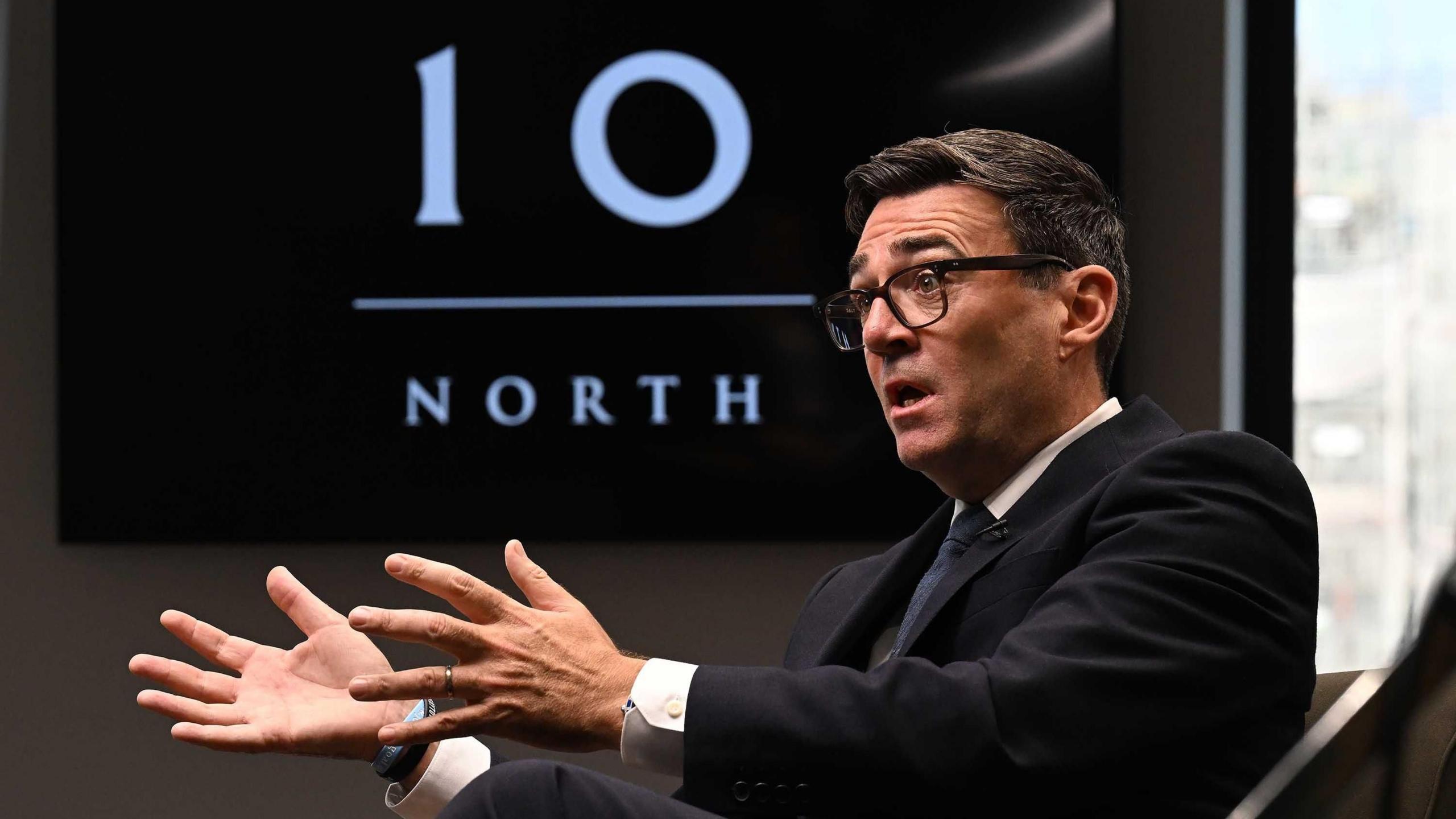 Andy Burnham, wearing a dark suit, gestures with his hands while speaking during an interview. Behind him is a TV with the logo for No 10 North.