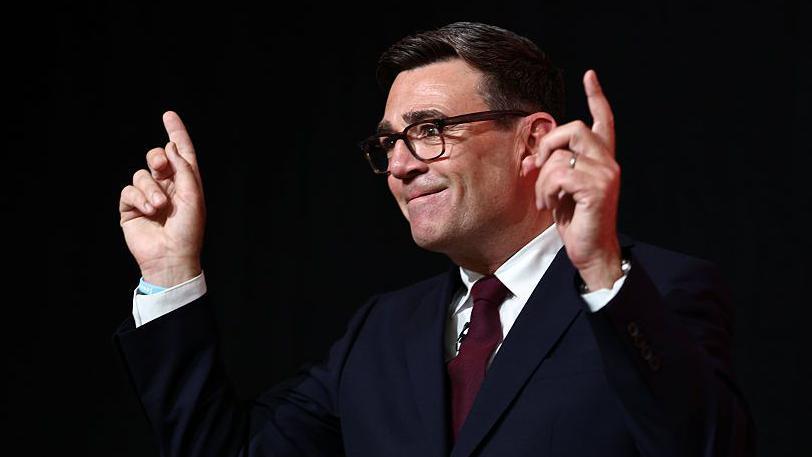 Andy Burnham, in a dark suit and red tie, raises both hands to point up, as he delivers his leadership 