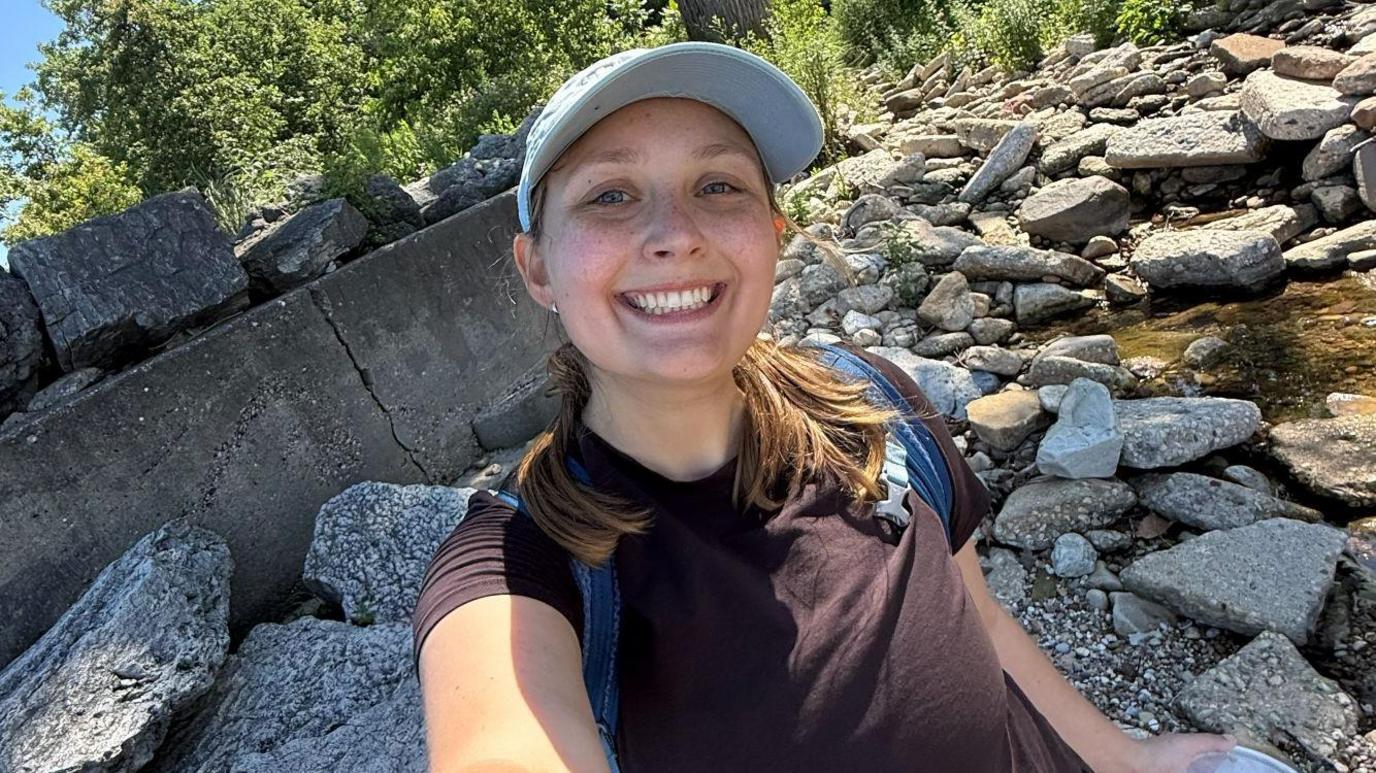 Megan Szostak stands in a rocky stream, taking a selfie and smiling at the camera. She wears a black t-shirt, blue backpack and blue hat.
