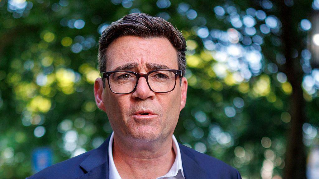 Andy Burnham wearing a white shirt and blue blazer. He is looking at the camera, and is wearing glasses.