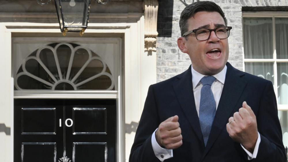 Andy Burnham is wearing a dark-coloured suit jacket, a white shirt and blue tie. He is also wearing glasses. He has black-coloured short hair. He is stood in Downing Street making a speech. Behind him is a black door with the number 10 on it.
