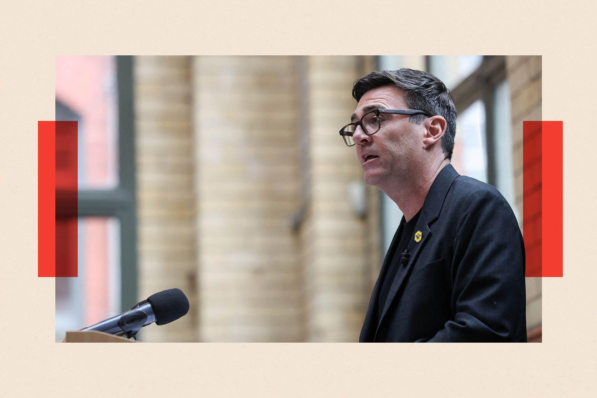 Andy Burnham delivers a speech at the People's History Museum in Manchester. He is viewed in profile standing at a podium. In the background are cream tiles and windows with green frames. Burnham has dark greying hair and wears dark-rimmed glasses and a dark jacket with a bee logo pin badge on his lapel.