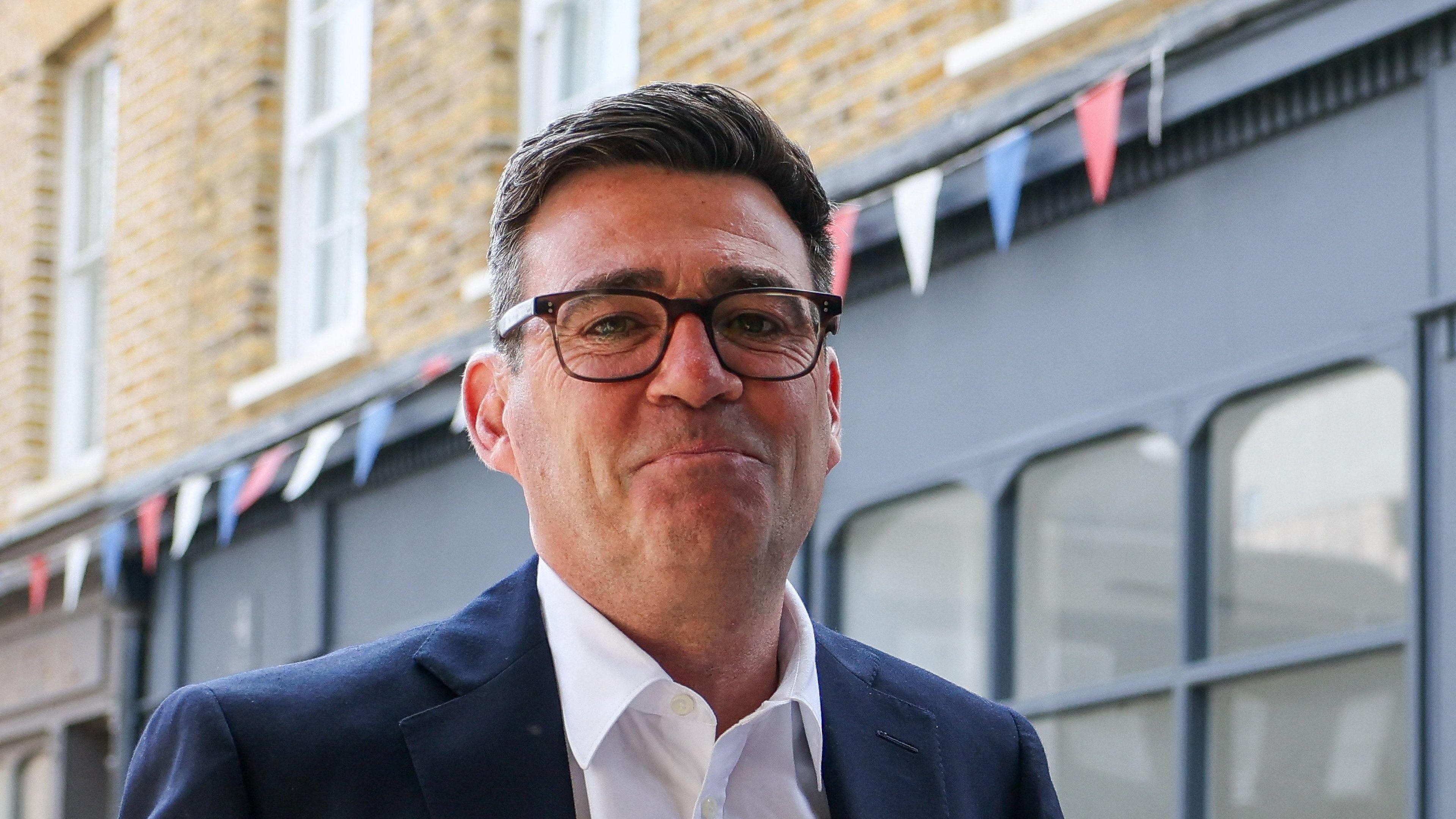 Andy Burnham looks directly forward and smiles with his mouth closed. He wears tortoise shell glasses, a navy suit jacket is navy and a white shirt. He stands in a street, behind him is a blue shop, with red, blue and white bunting hung above it.