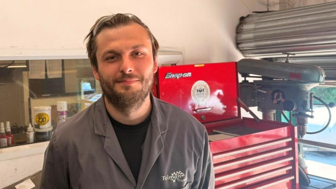 Ollie Priestley wearing a grey work overall stands in front of a red tool cabinet.