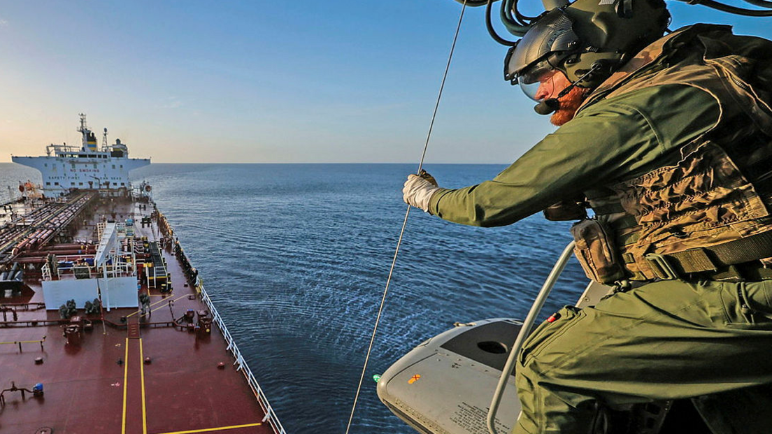 A still showing a military personnel on a helicopter above a large vessel. He is wearing green tactical gear and a helmet and gloves and is looking down at the boat. The water and sky are blue. 