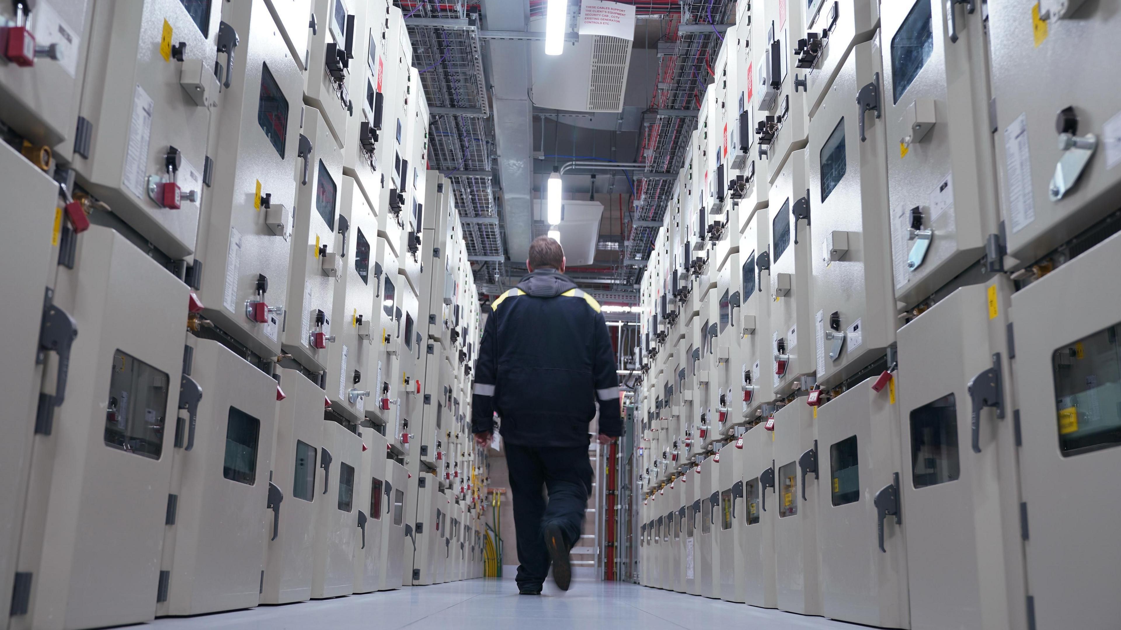 A man in dark jacket walks between two rows of beige coloured metal boxes under strip lighting in the high voltage switching room of a London data centre