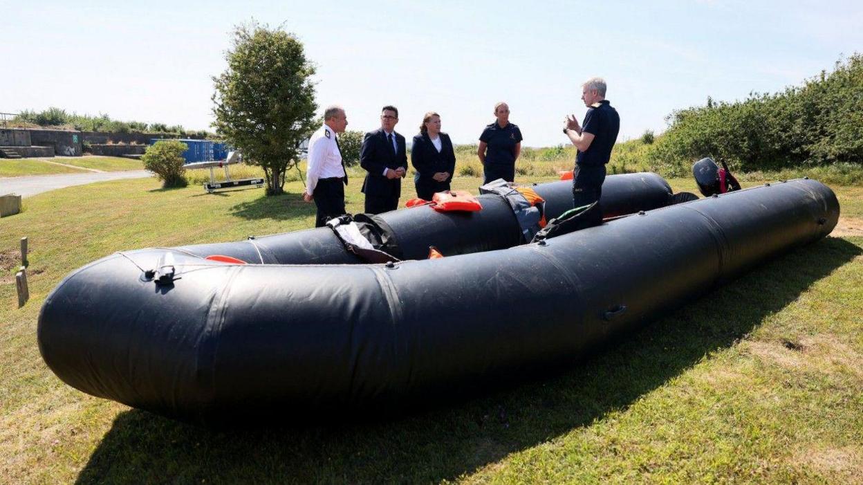 Wide shot showing Andy Burnham with his Minister for Border Security and Asylum Anna Turley and three officials standing by a recovered small boat. One official is standing inside the boat a black inflatable dinghy that is a few metres long.