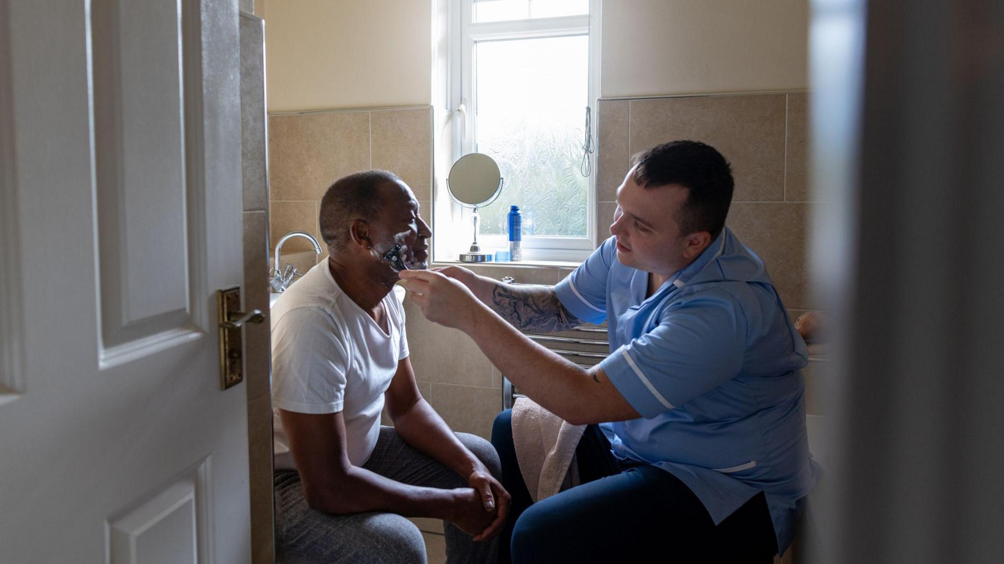 A care worker in a blue uniform helps a seated person shave in a bathroom. The two are positioned beside a window, with a sink, mirror and toiletries visible in the room.
