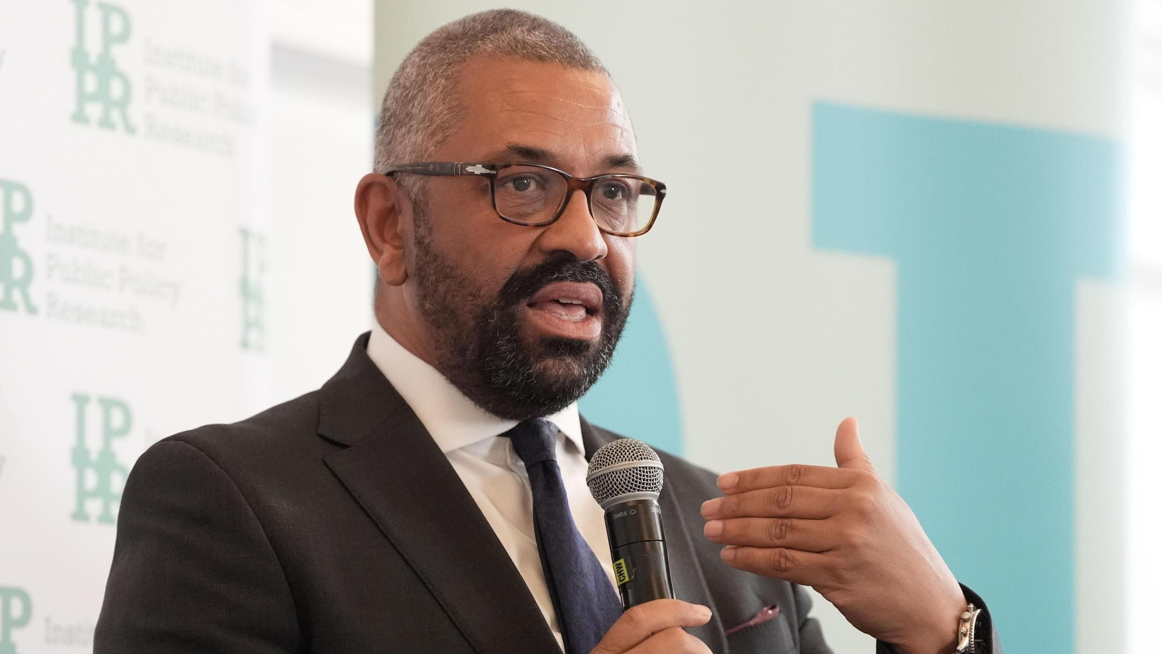 James Cleverly wears a suit and holds a microphone