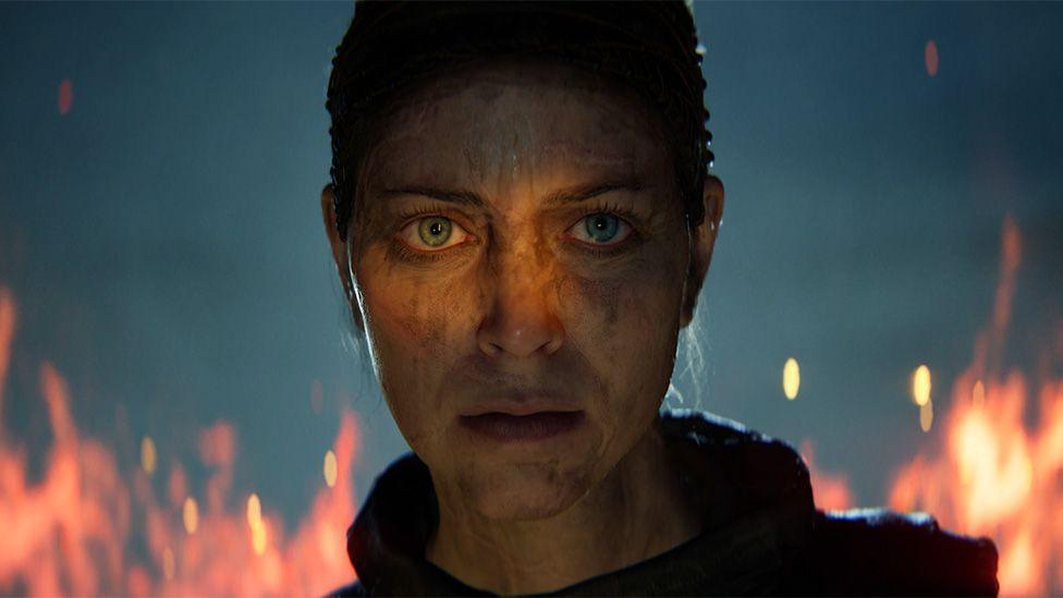 An in-game close-up of the female protagonist in Hellblade, with flames burning behind her