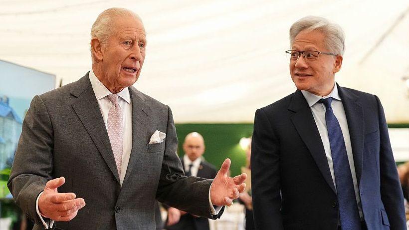 King Charles wearing a grey suit jacket and white shirt and pink tie, talking to Jensen Huang, the CEO of Nvidia, who is wearing a dark suit, white shirt and blue tie. 