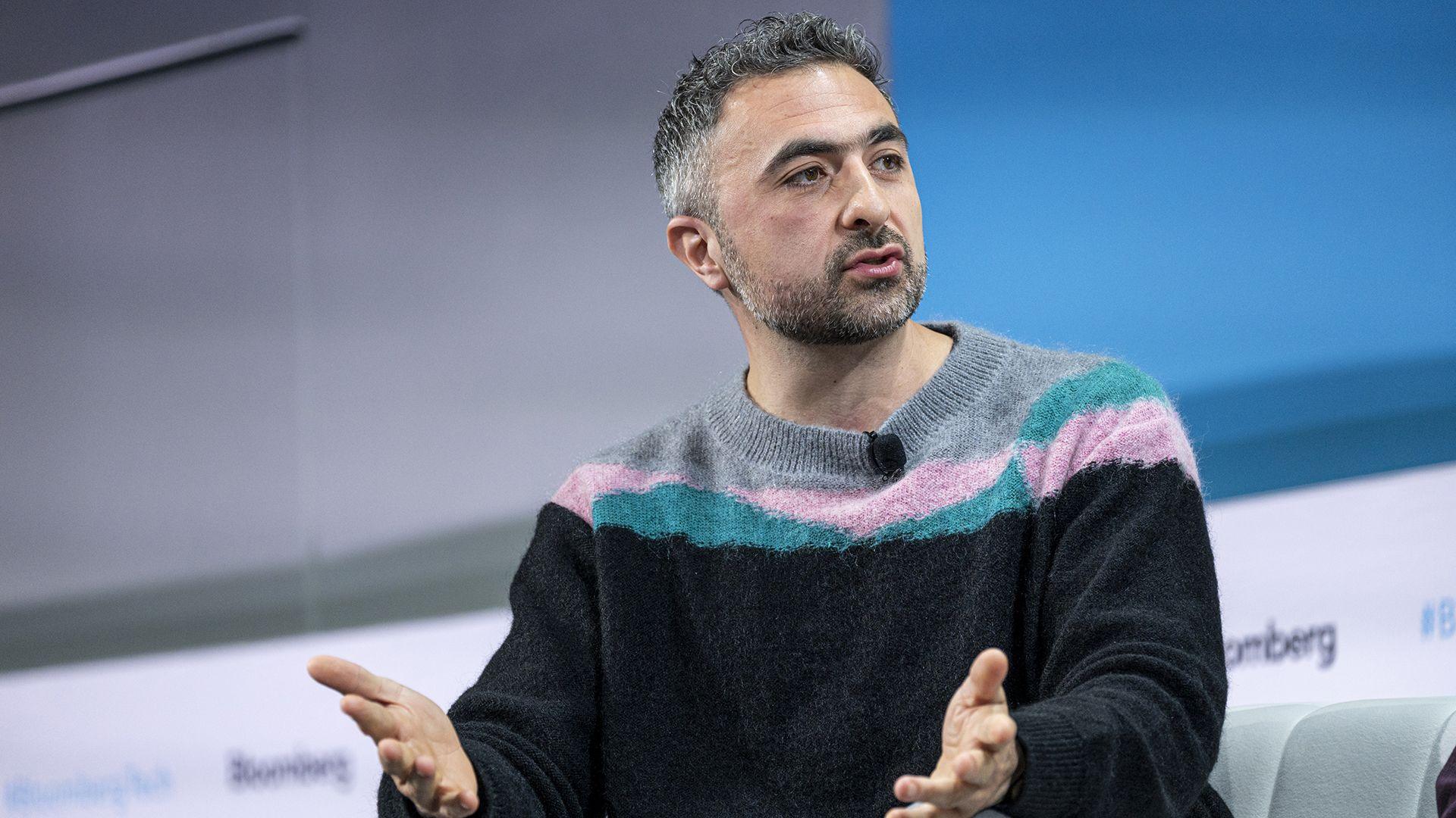 Mustafa Suleyman sitting on a sofa at a conference in the middle of speaking, wearing a black, turquoise and pink jumper