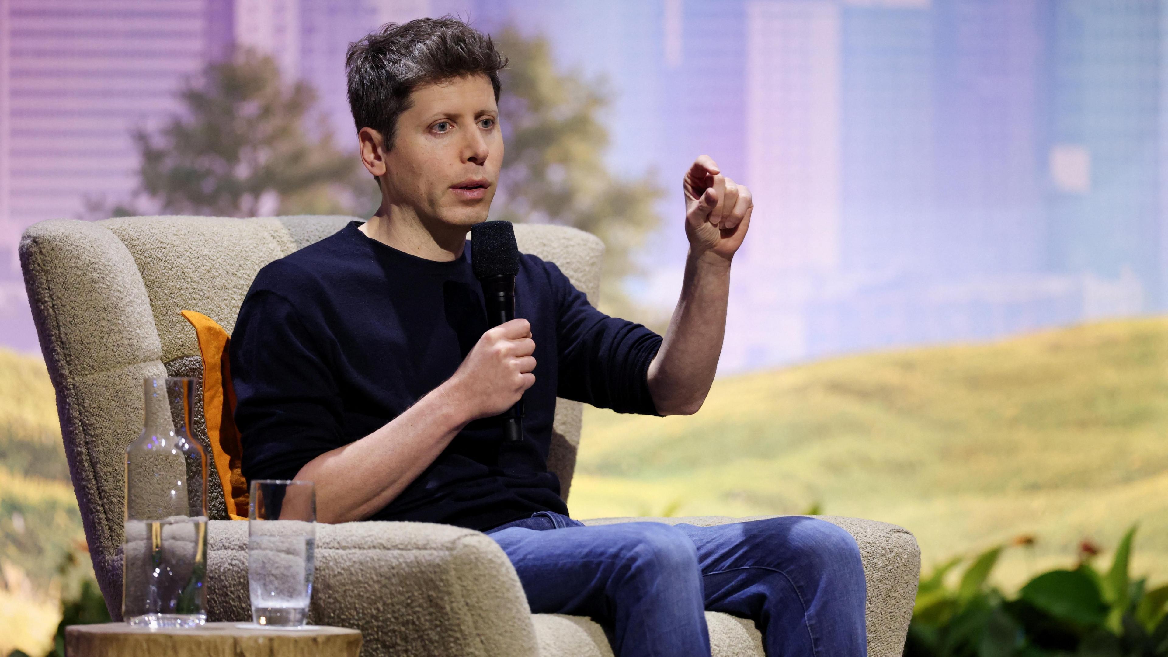 Sam Altman sitting on stage at the conference in Washington, gesturing with his left hand 