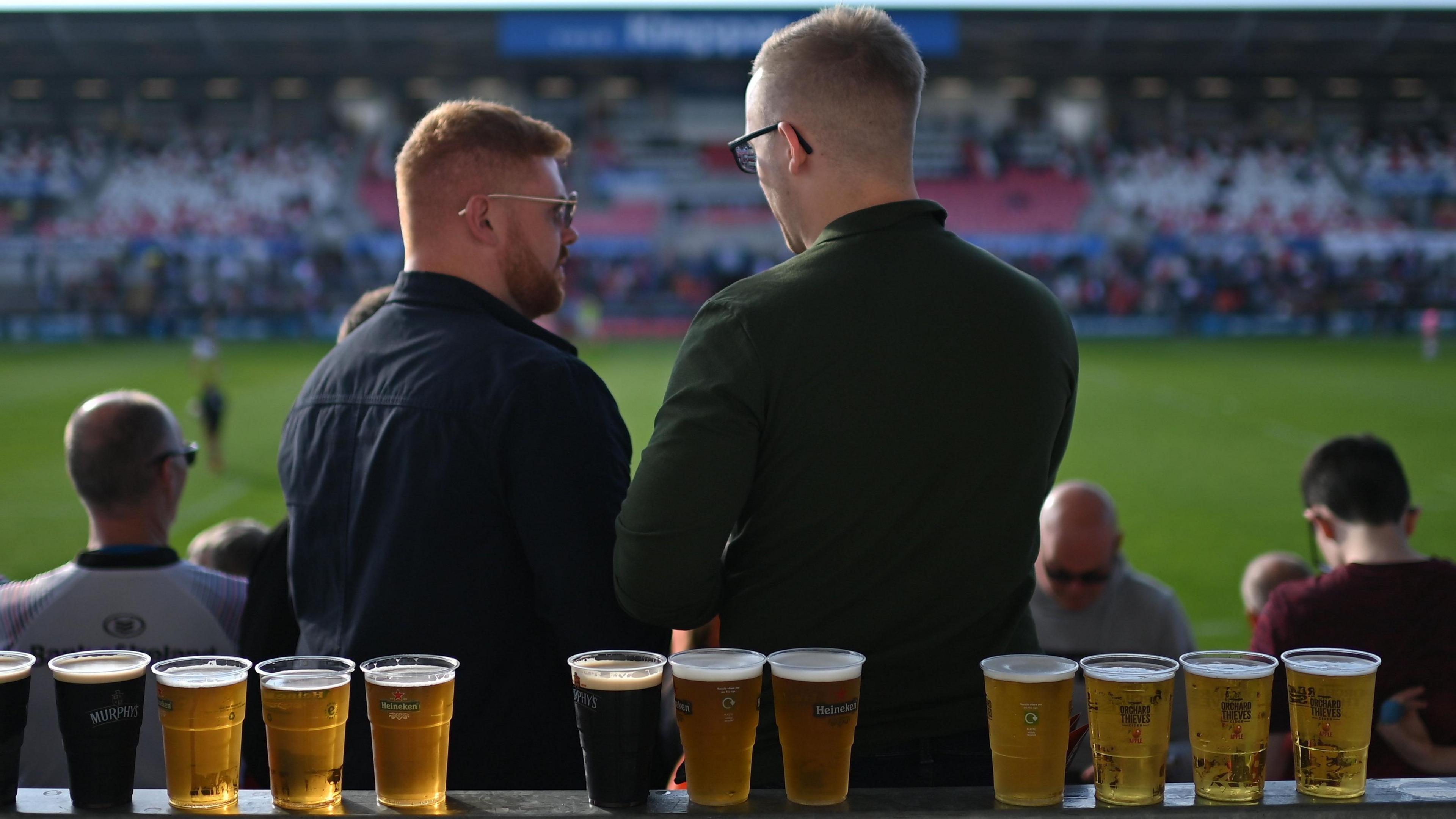 Two men at a sporting event stand in front of a row of pints of beer along a rail.