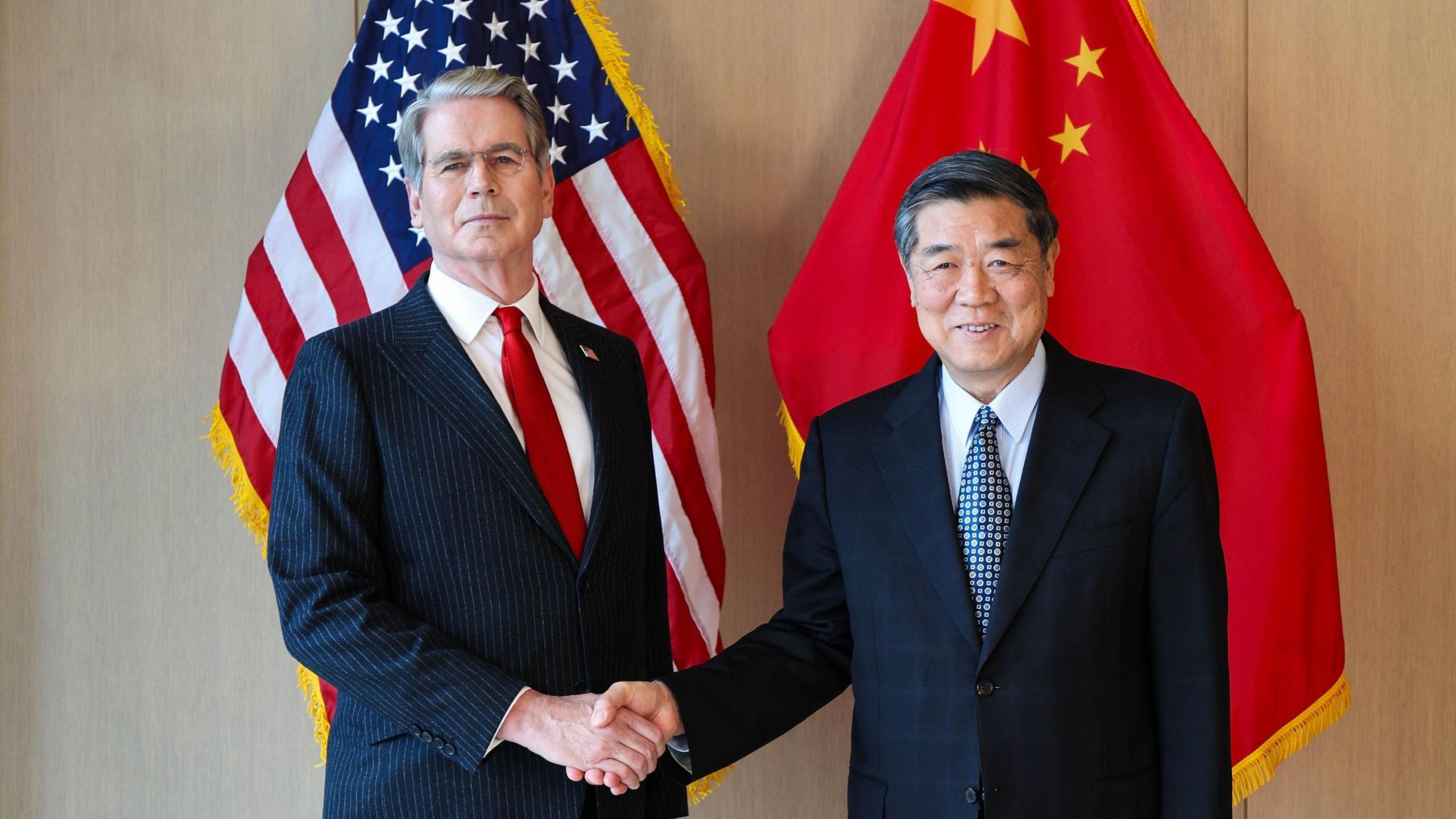 US Treasury Secretary Scott Bessent shakes the hand of China's top trade negotiator He Lifeng stand in front of both countries' flags.