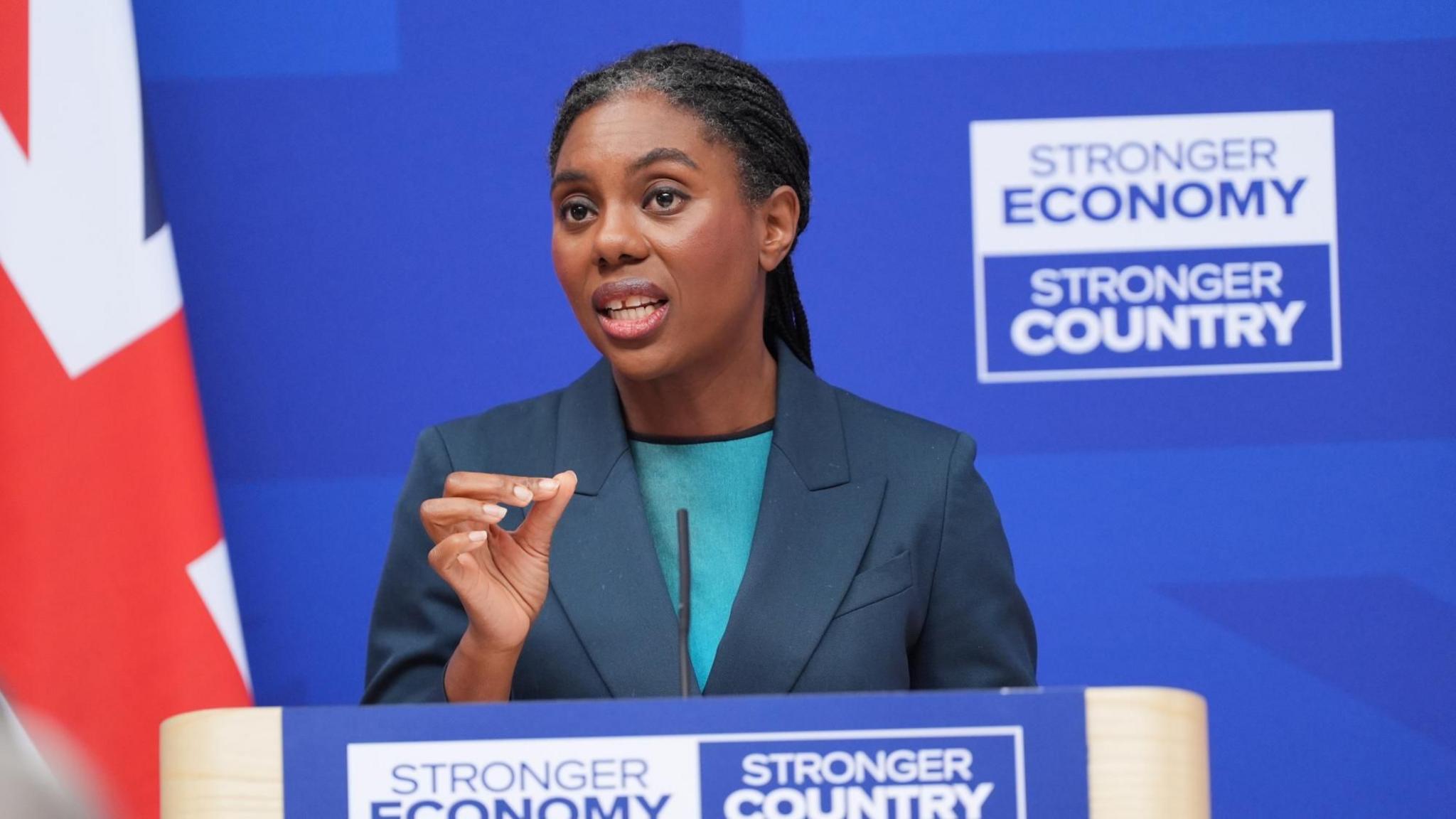 Badenoch is pictured from below the podium where she is giving a speech, at London Scottish House in Westminster, with a royal blue background and a Union flag to her left. She wears her braided hair tied back in a low ponytail, with a navy jacket and a turquoise top.